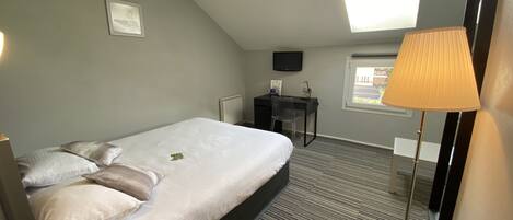 Habitación de hotel con cama, escritorio, silla, televisor, ventana y lámpara.