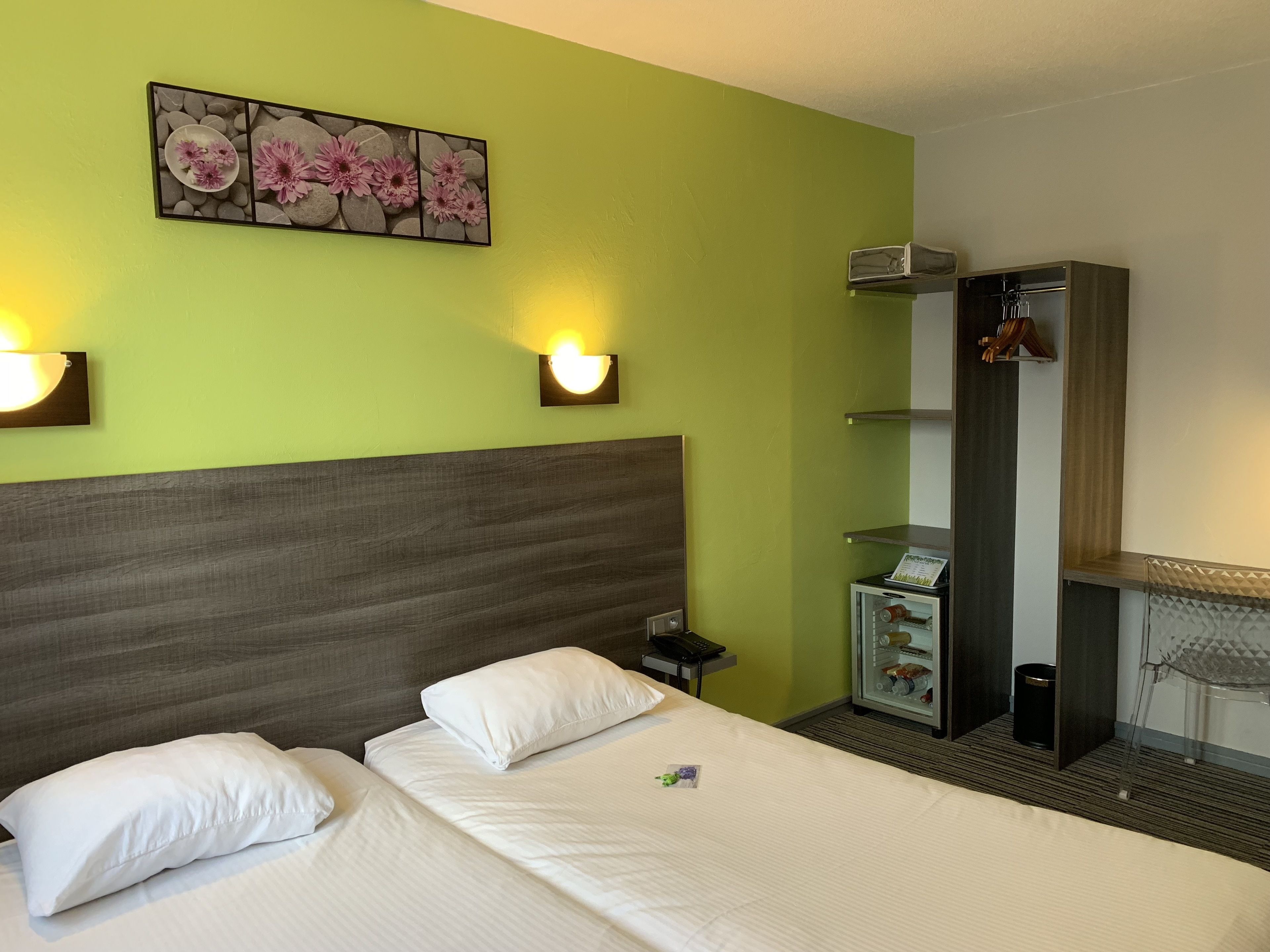 A hotel room with a bed, a nightstand, a wall-mounted light, a small table with a phone, and a shelf with a mirror and a small cabinet.