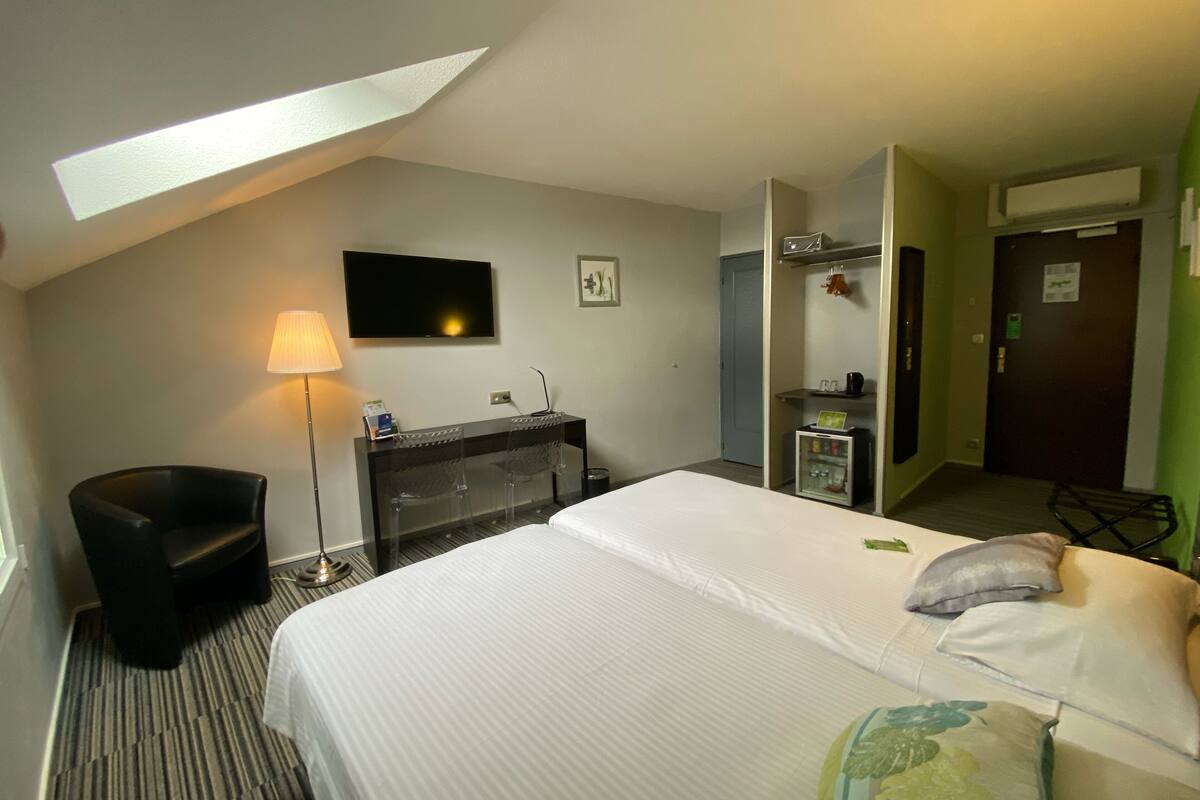 Quarto de hotel com cama, escrivaninha, cadeira, televisão, candeeiro e minibar.