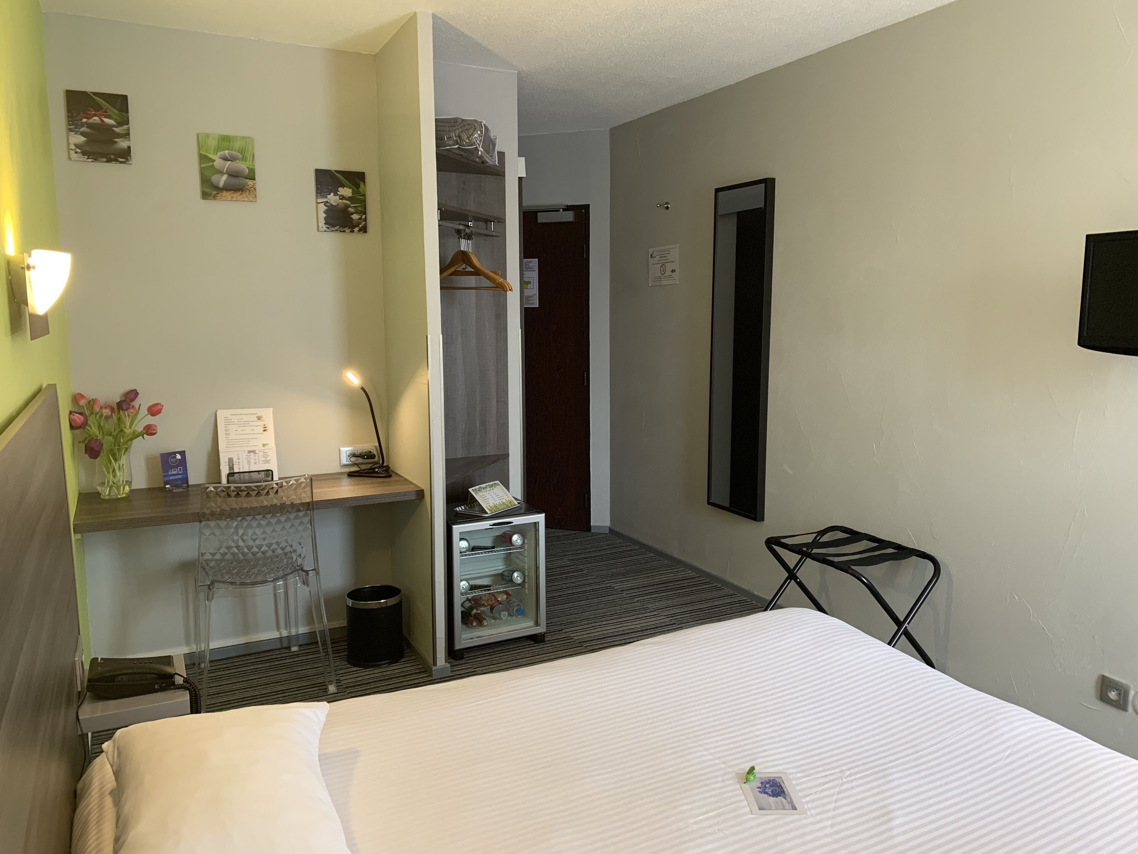 Habitación de hotel con una cama, un escritorio con lámpara, un espejo y un pequeño estante con un florero.