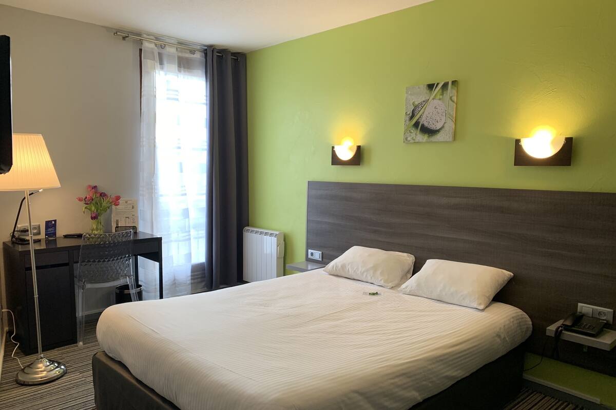 Quarto de hotel com uma cama grande, uma escrivaninha com luminária, uma cadeira, radiador e duas luzes na parede.