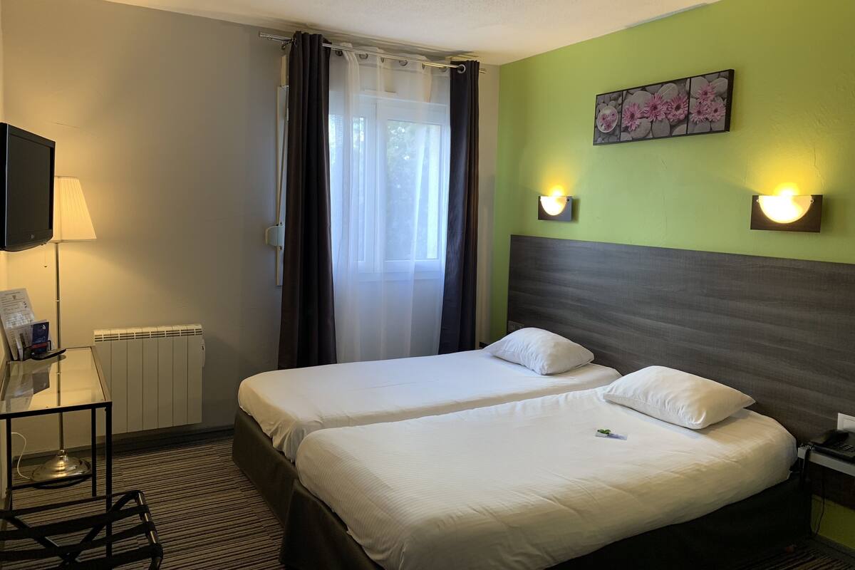 Quarto de hotel com duas camas, televisão, candeeiro, radiador e janela com cortinas.