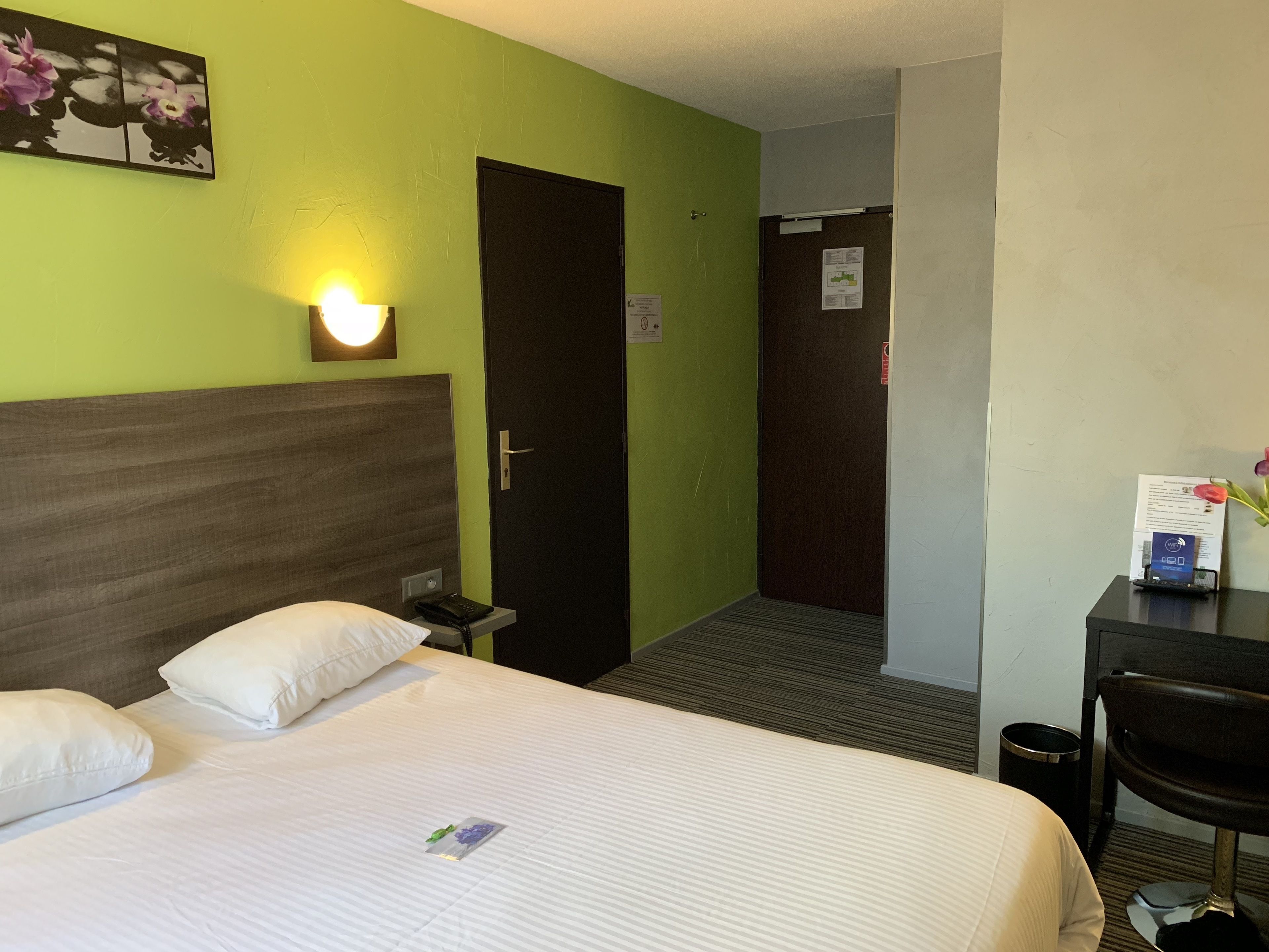 Hotellihuone, jossa on sänky, työpöytä ja tuoli. Taustalla on ovi ja seinässä valaisin.