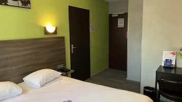 Une chambre d’hôtel avec un lit, un bureau et une chaise. On aperçoit une porte au fond et un mur avec un luminaire.