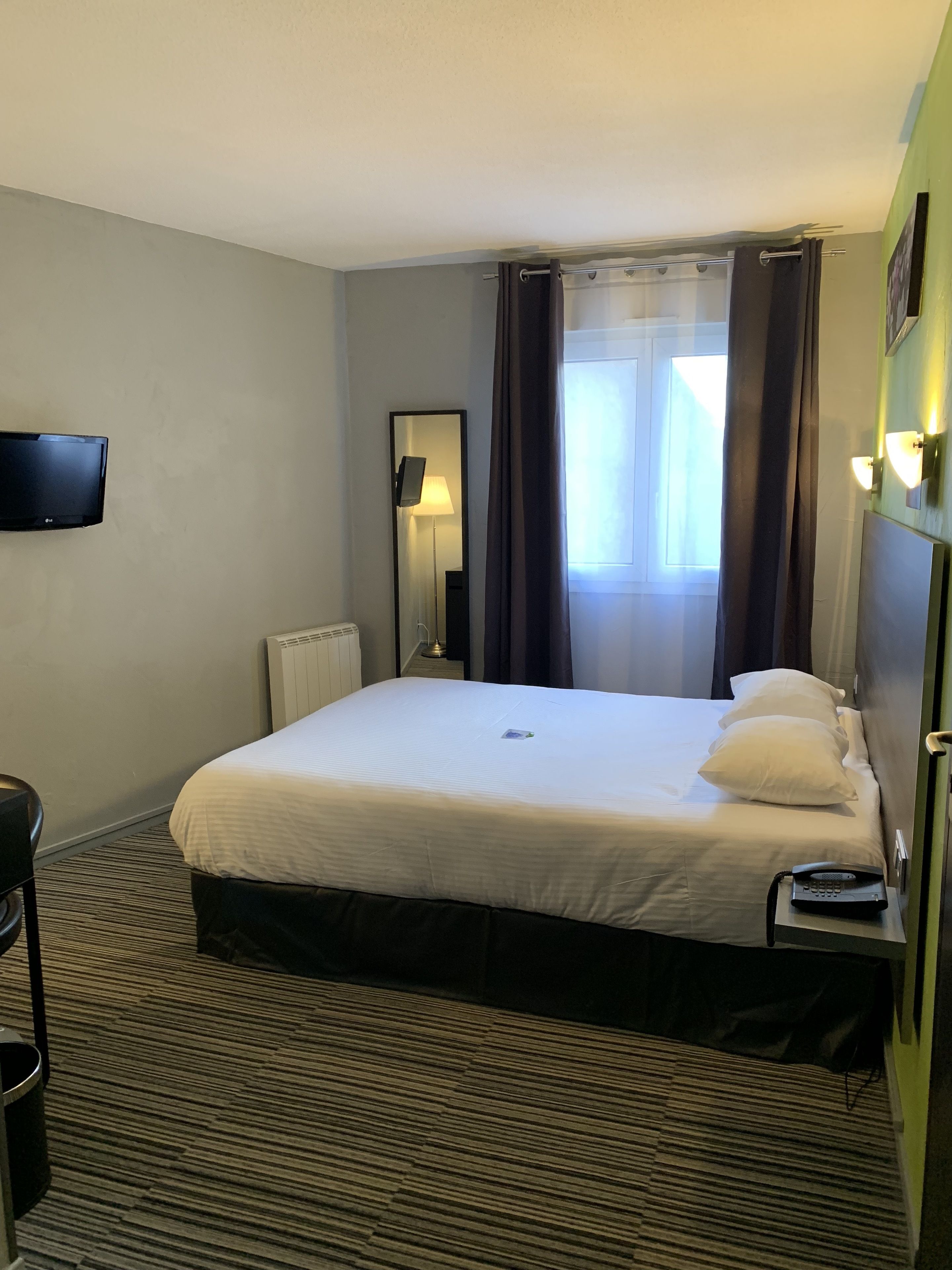 Habitación de hotel con cama, mesita de noche con teléfono, televisor de pantalla plana montado en la pared, radiador y ventana con cortinas.