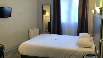 Habitación de hotel con cama, mesita de noche con teléfono, televisor de pantalla plana montado en la pared, radiador y ventana con cortinas.