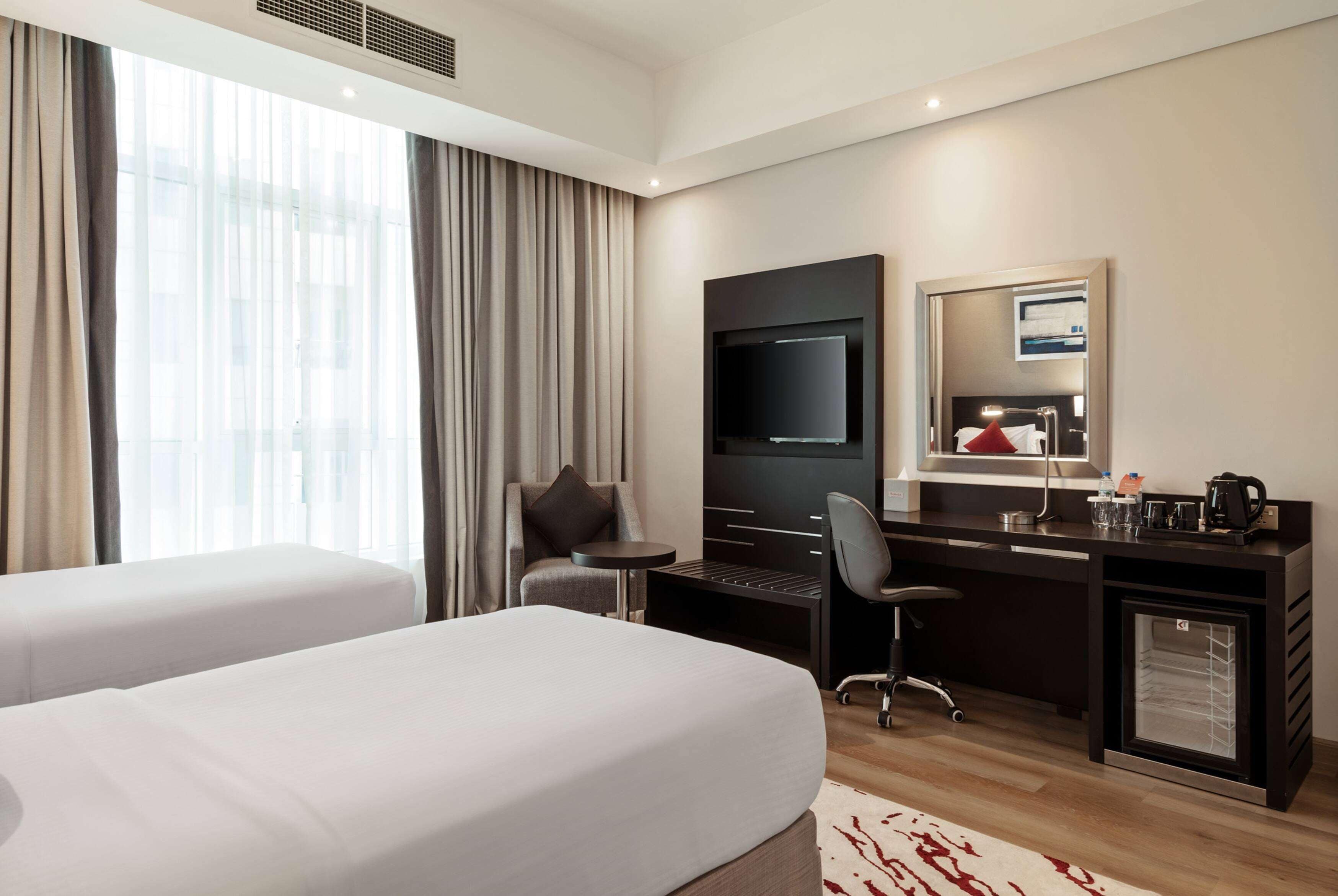 A hotel room with two beds, a desk, a chair, a television, and a minibar.