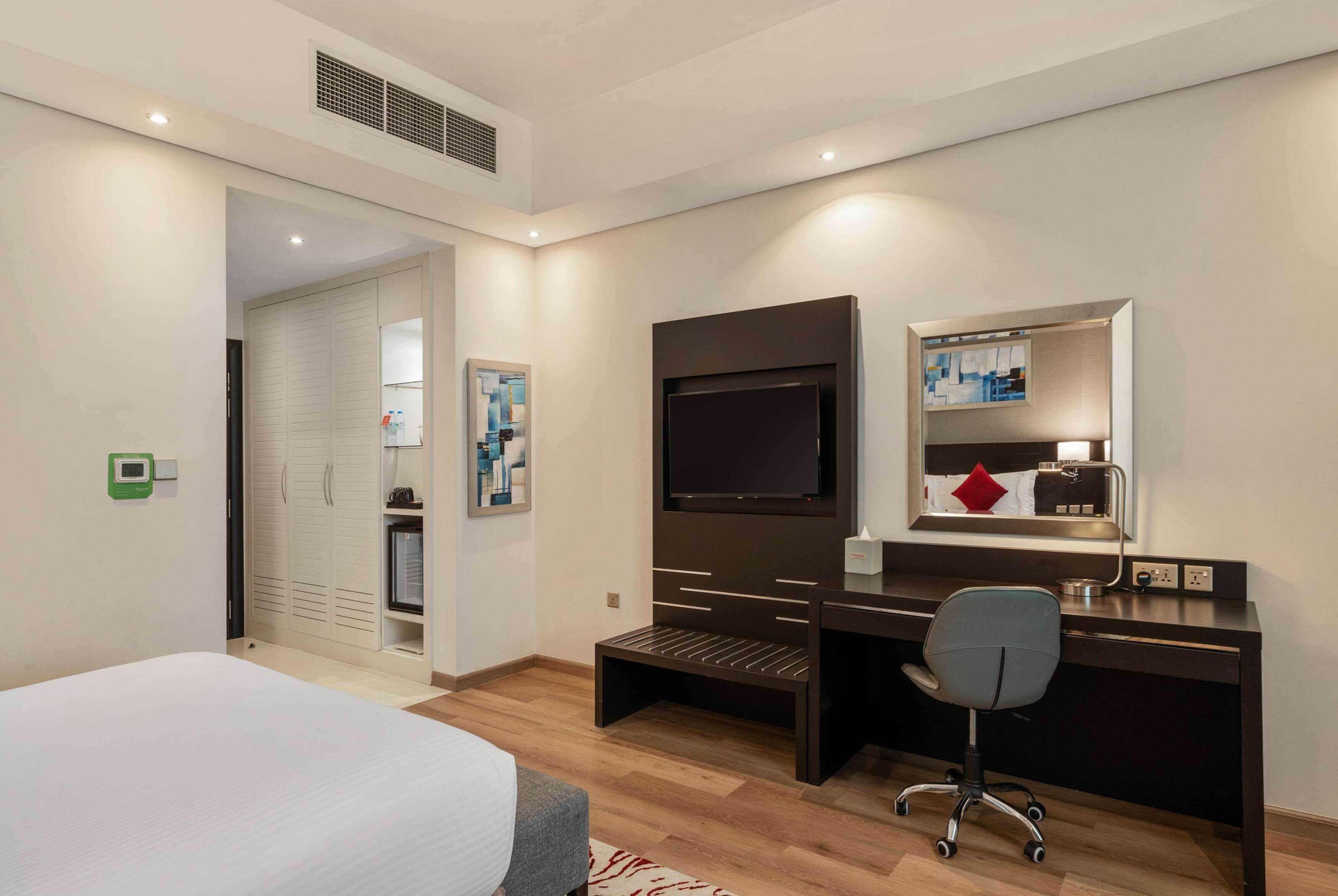 A hotel room with a bed, desk, chair, TV, wardrobe, and a small shelf with toiletries.