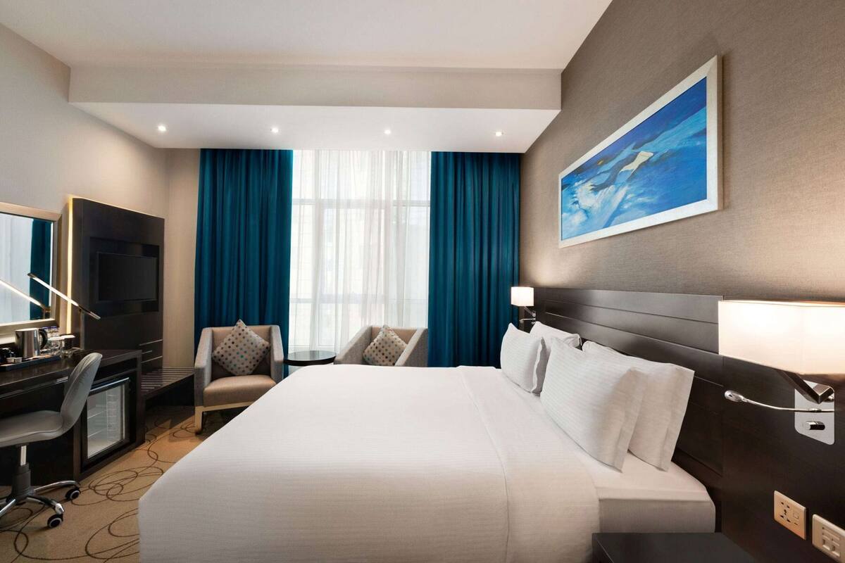 A hotel room with a bed, two armchairs, a desk with a TV, and a painting on the wall.