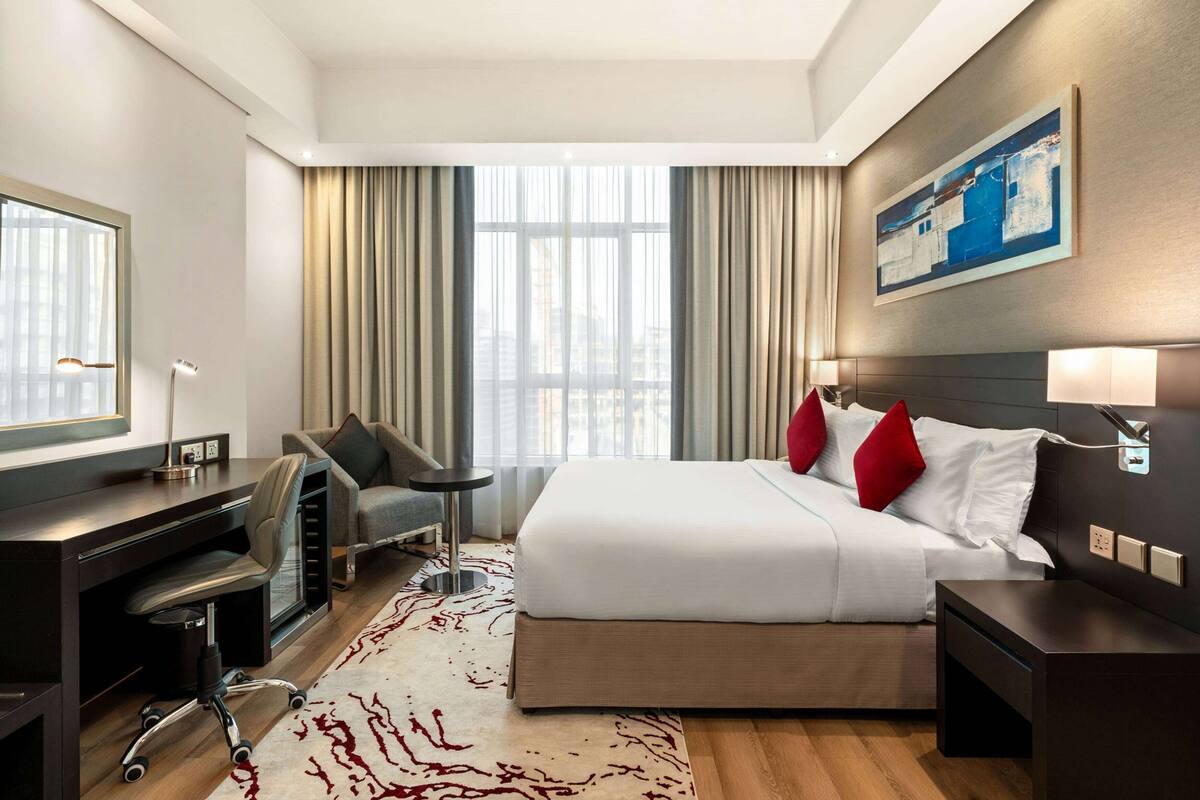 A hotel room with a bed, desk, chair, and a large window with curtains.