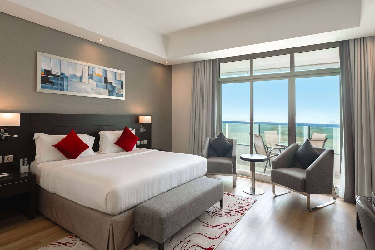 A modern hotel room with a large bed, a bench, a chair, and a view of the sea.