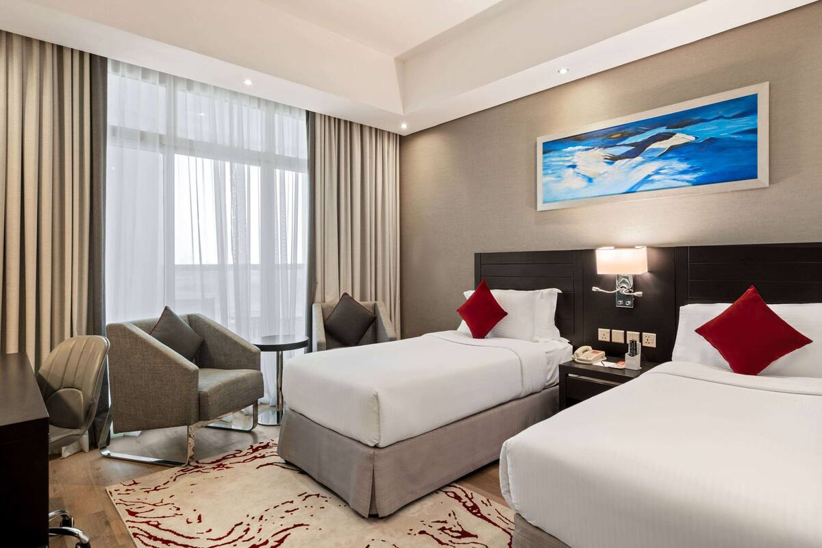 A hotel room with two beds, a desk, a chair, and a large window with curtains.