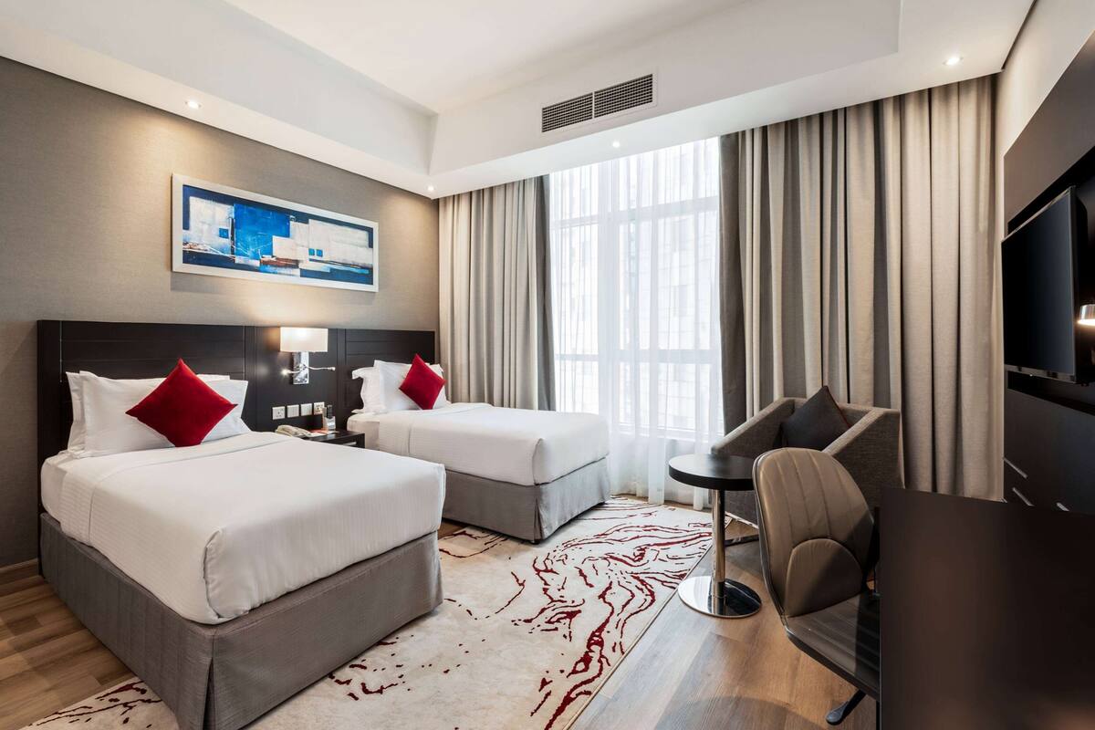 A hotel room with two beds, a desk, a chair, a TV, and a large window with curtains.