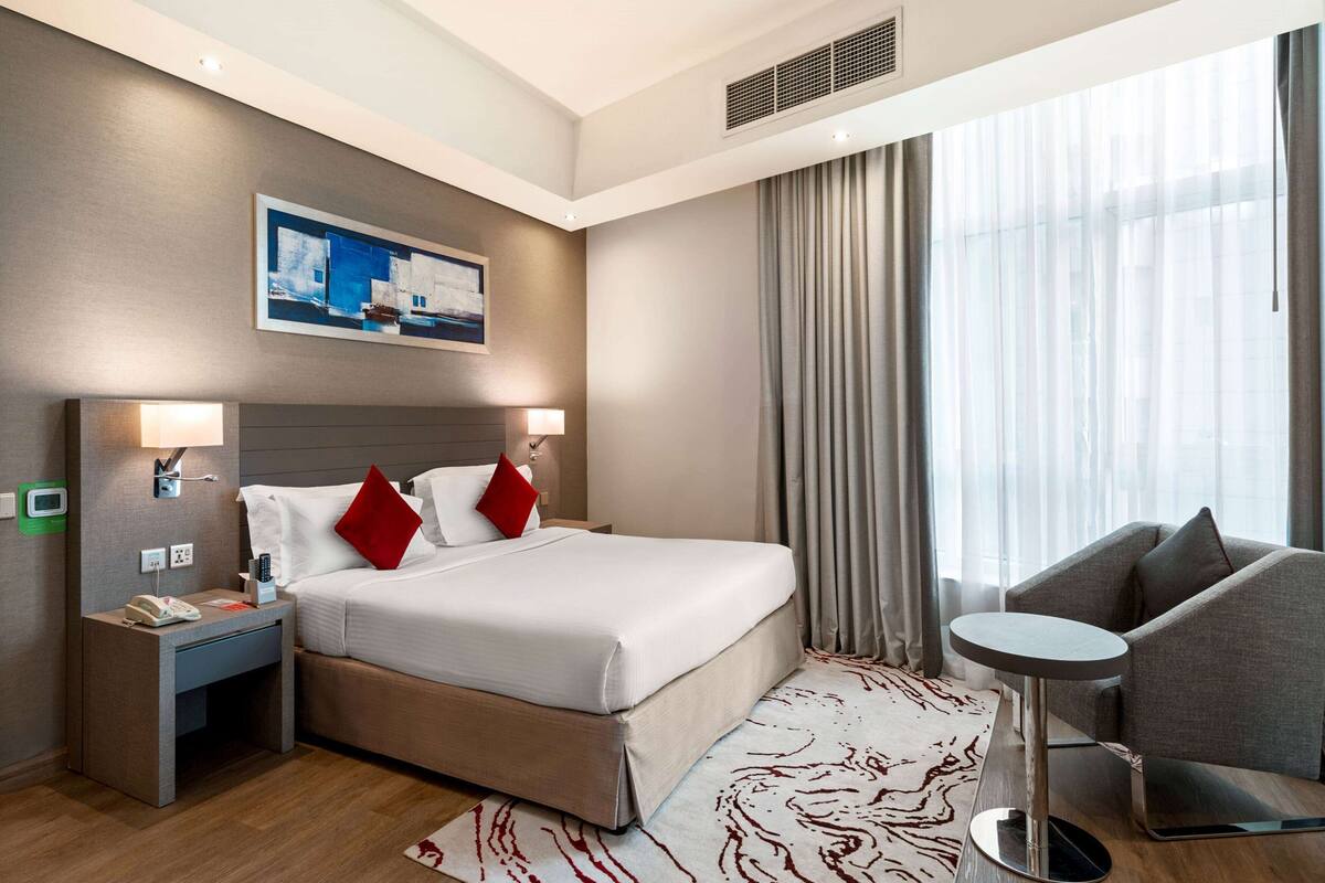 A hotel room with a large bed, bedside lamps, a small table, and a view of the outdoors through sheer curtains.
