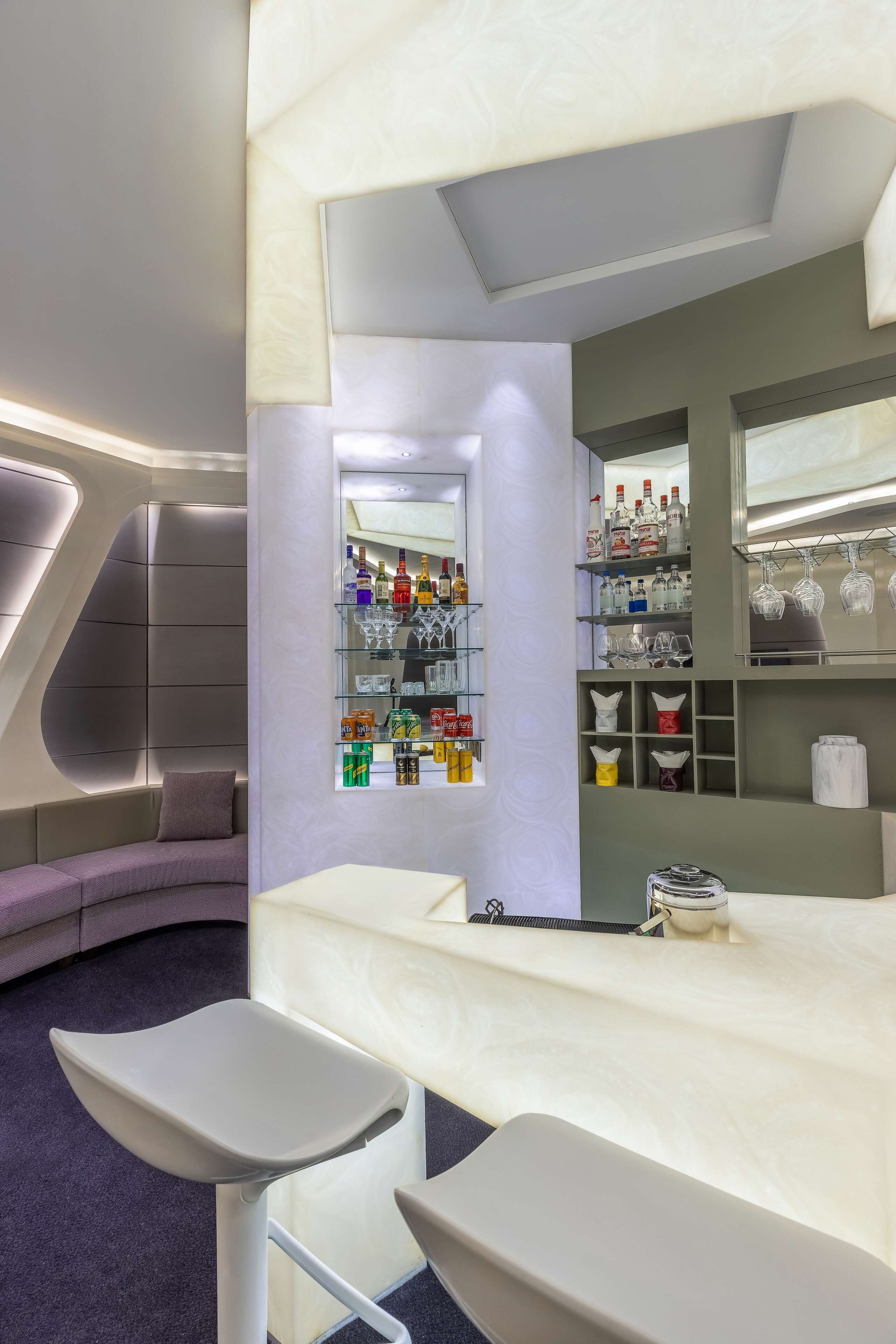 A modern bar area with a curved seating arrangement, a minibar, and shelves stocked with bottles and glasses.