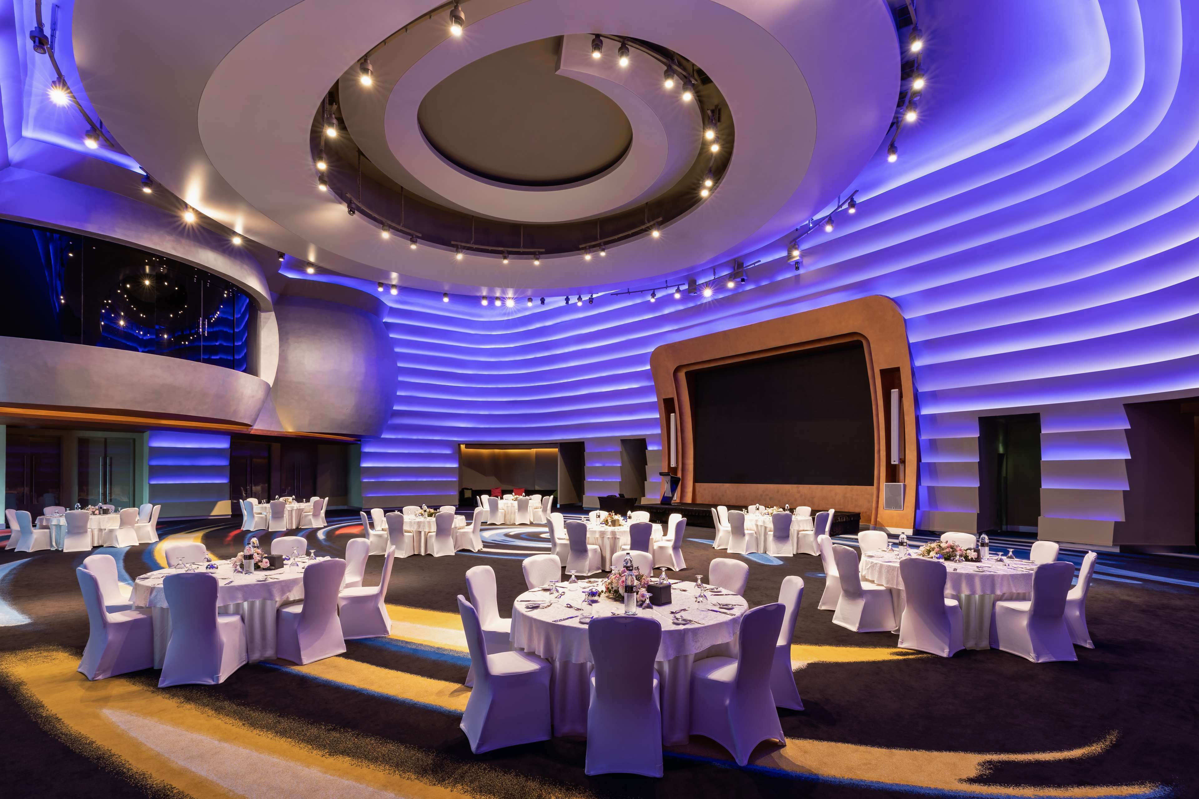 A modern conference room with round tables and white chairs, a large screen, and a circular ceiling design.