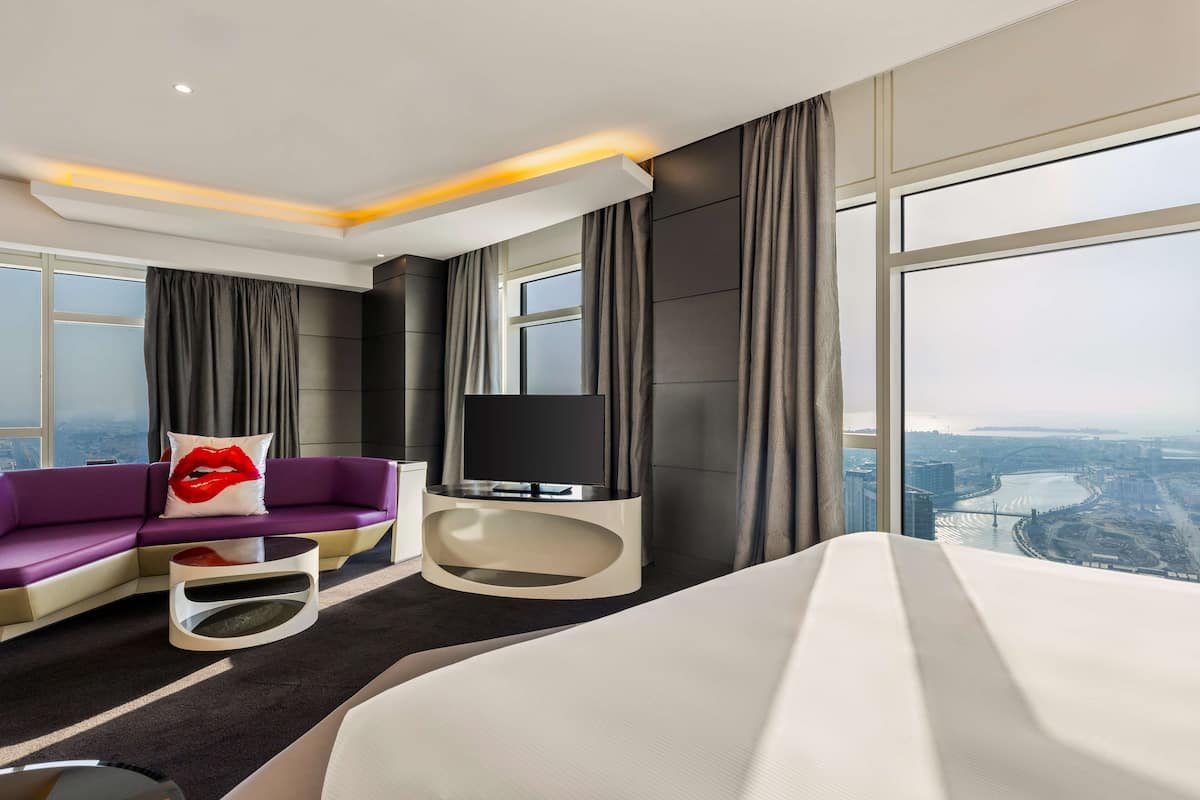 Una moderna camera d'albergo con un letto grande, un divano, una TV e vista sulla città attraverso ampie finestre.