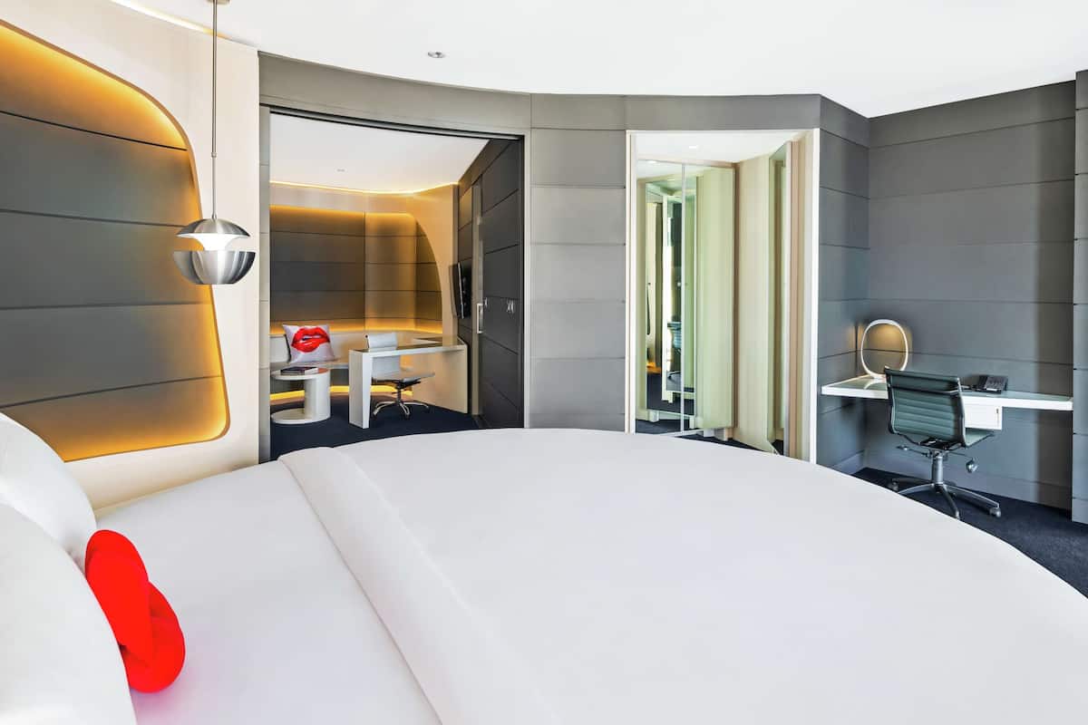 Una camera d'albergo moderna con un letto grande, una scrivania, una sedia, uno specchio e un bagno visibile attraverso una porta aperta.