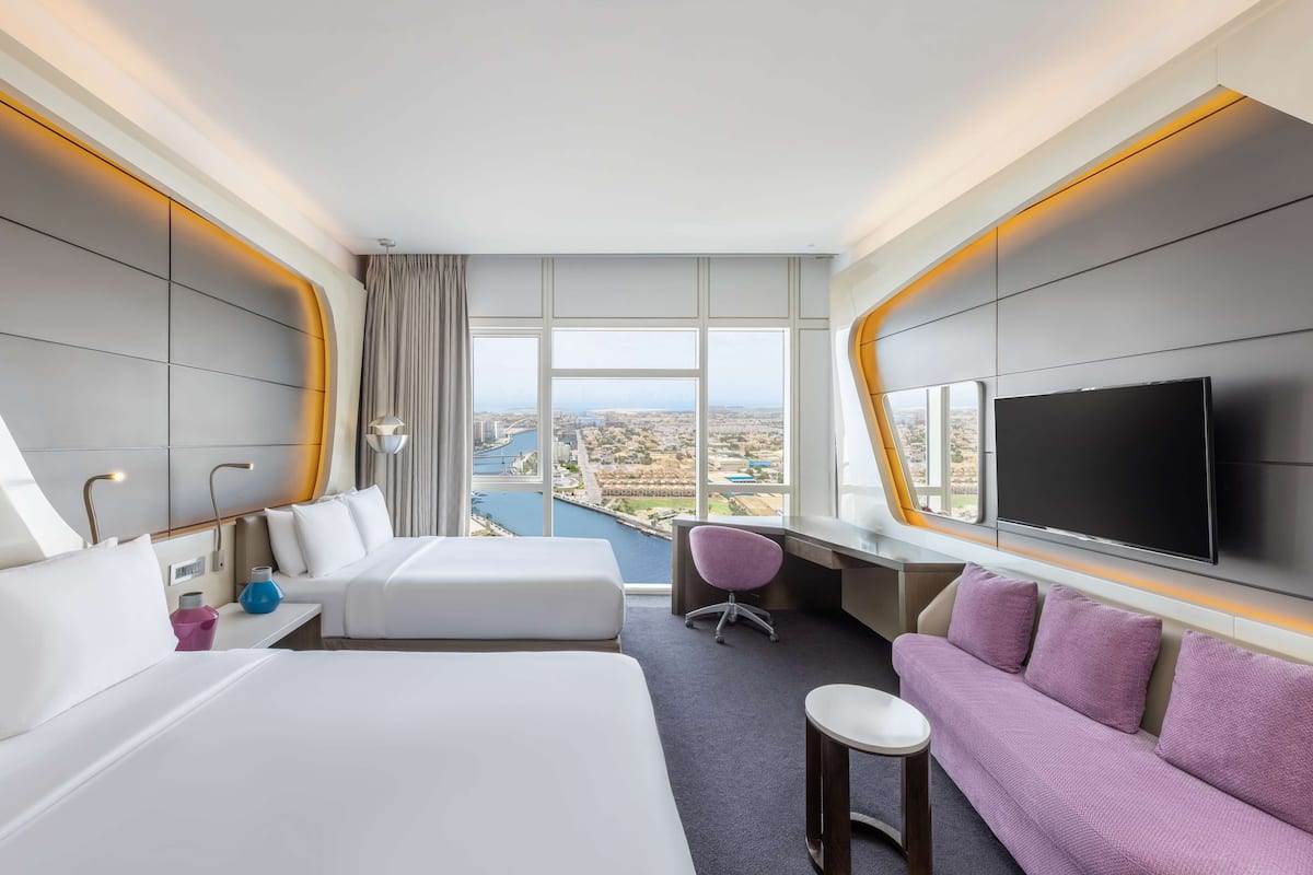 Una moderna camera d'albergo con un letto grande, un divano, una scrivania e una televisione. La stanza ha ampie finestre che offrono vista su un panorama urbano e uno specchio d'acqua.