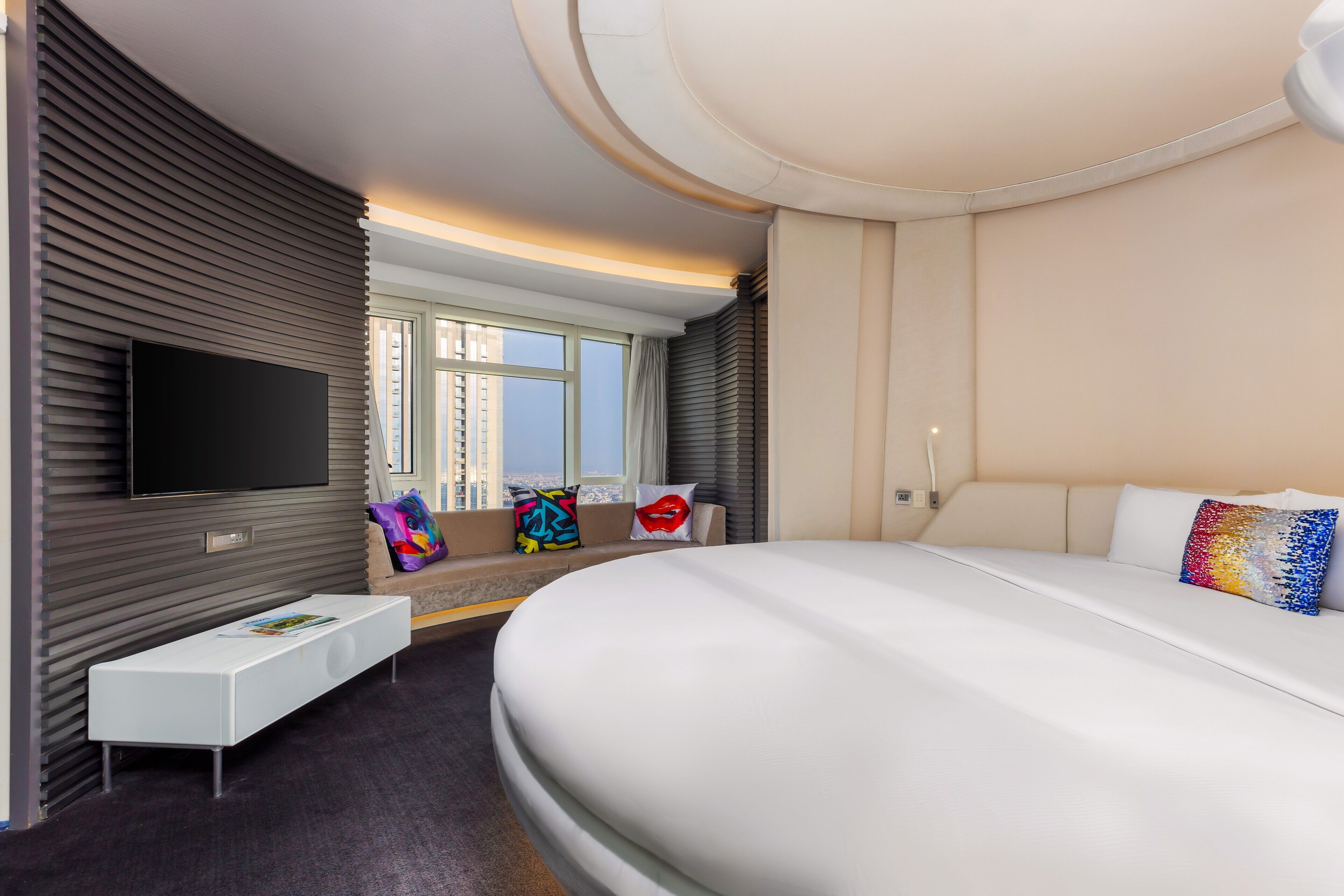 A modern hotel room with a large bed, a flat-screen TV mounted on a wall, a small white table, and a window with a view of the city.