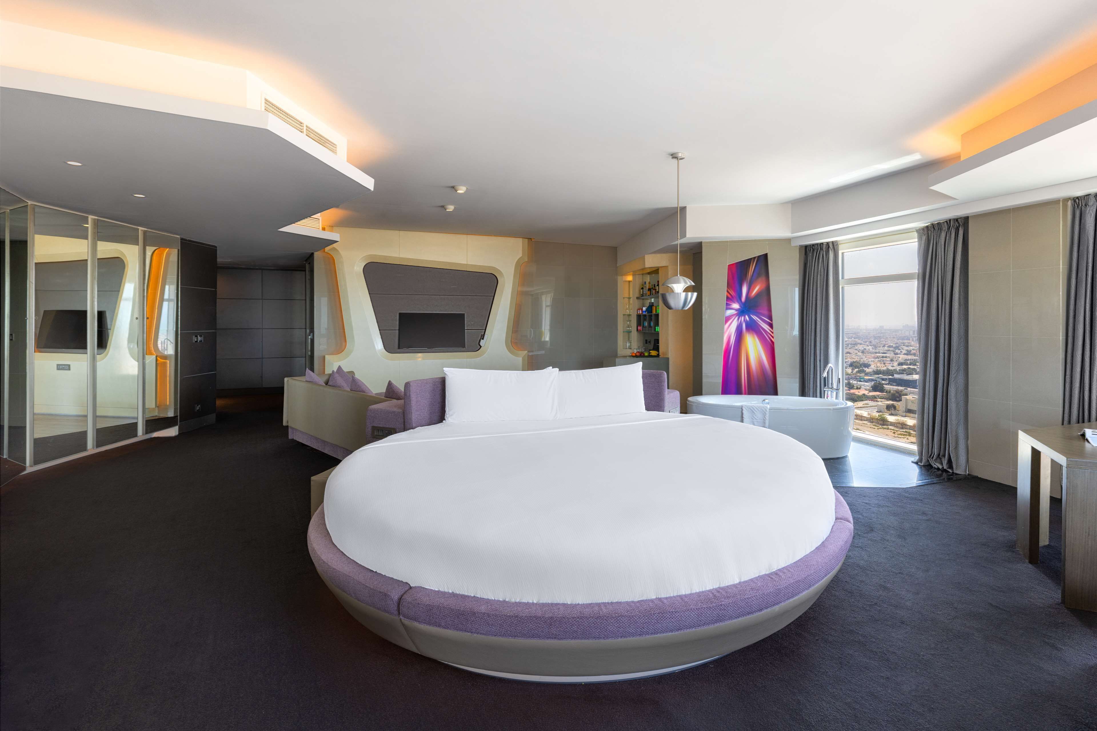 A modern hotel room with a large bed, a TV, a seating area, and a view of the city.