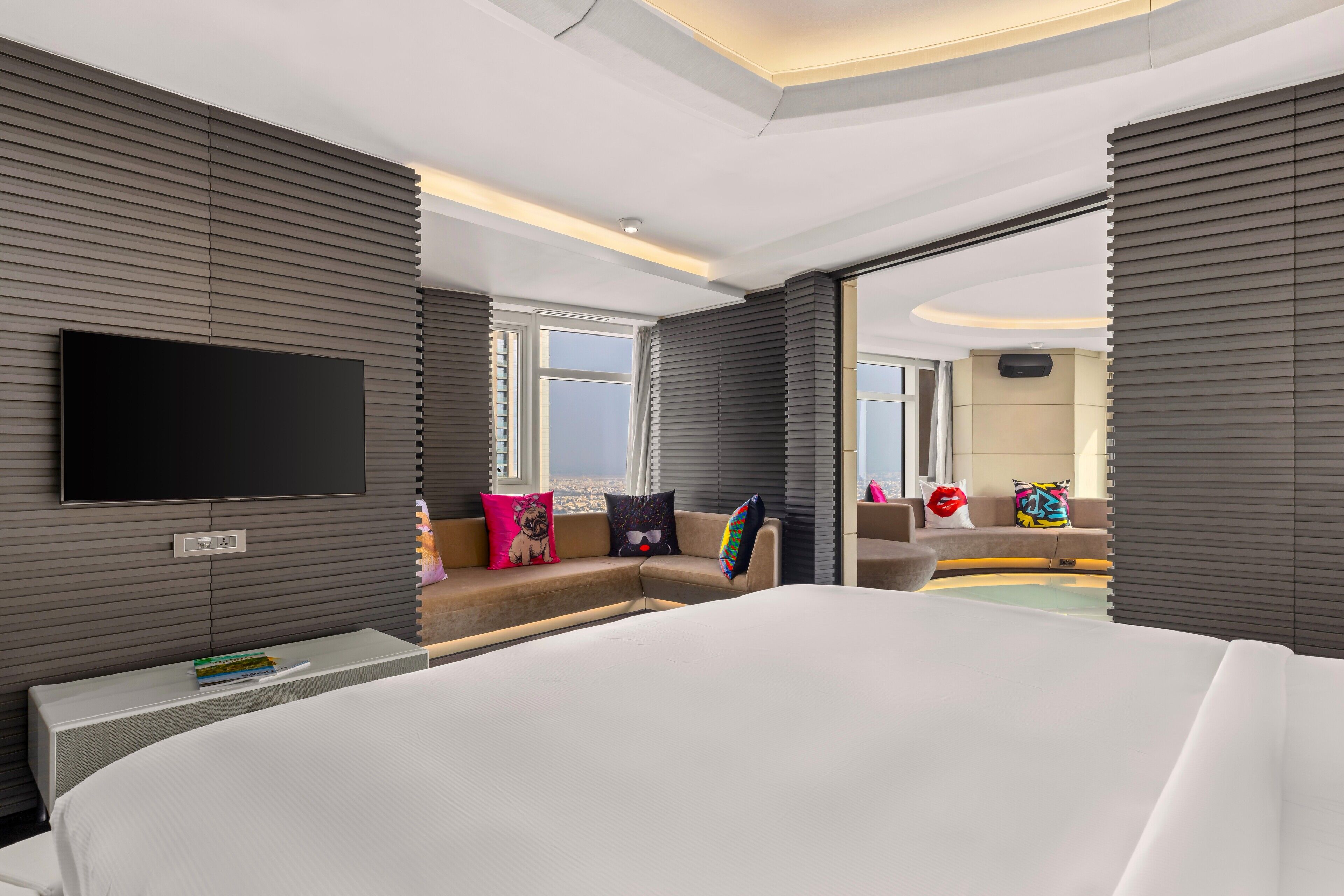 A modern hotel room with a large bed, a flat-screen TV mounted on the wall, a sofa with colorful cushions, and a view of the city through the window.
