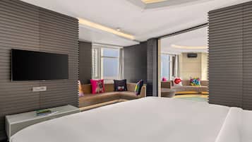 A modern hotel room with a large bed, a flat-screen TV mounted on the wall, a sofa with colorful cushions, and a view of the city through the window.