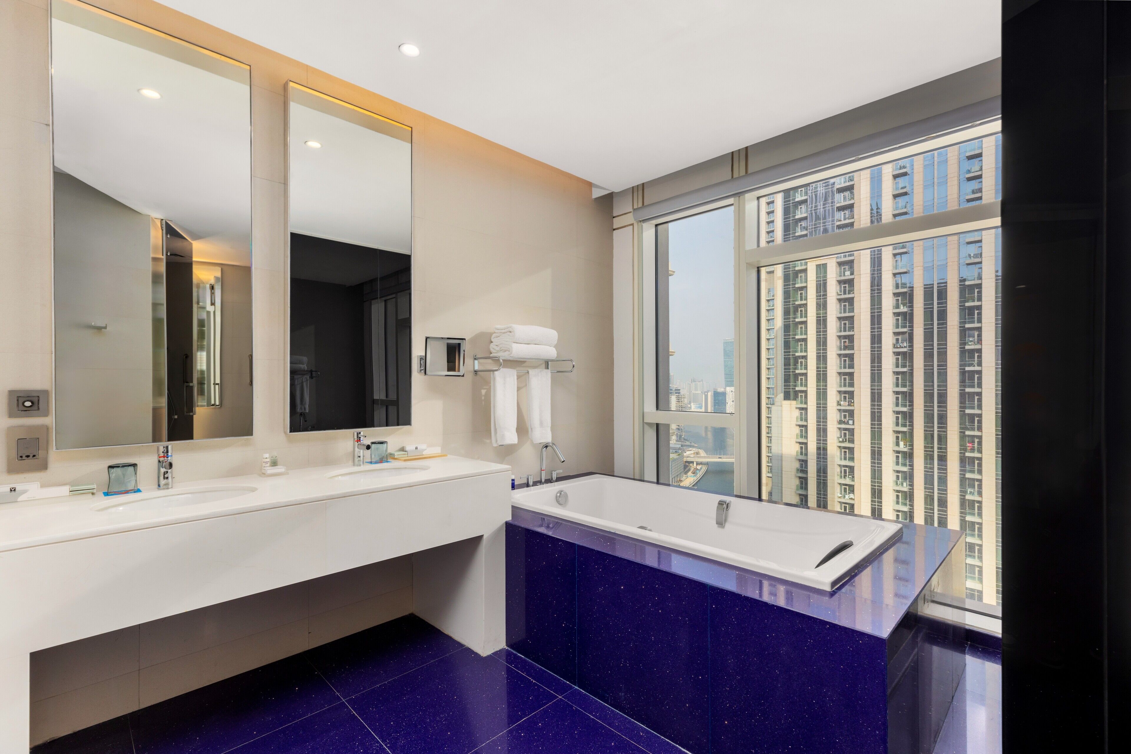A modern bathroom with a large mirror, two sinks, a bathtub, and a view of tall buildings through a large window.