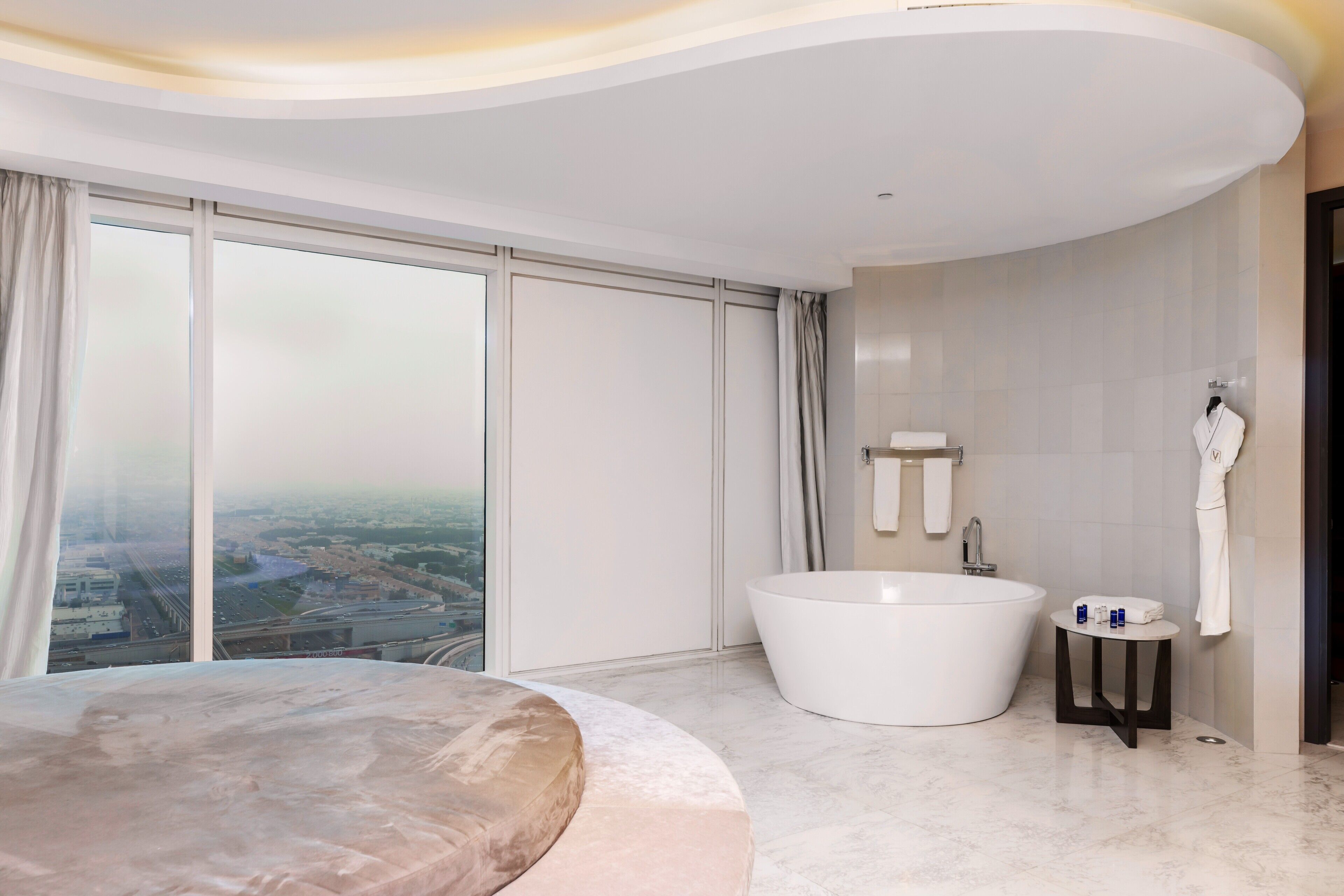 A modern hotel room with a large bed, a freestanding bathtub, and a view of a cityscape through large windows.