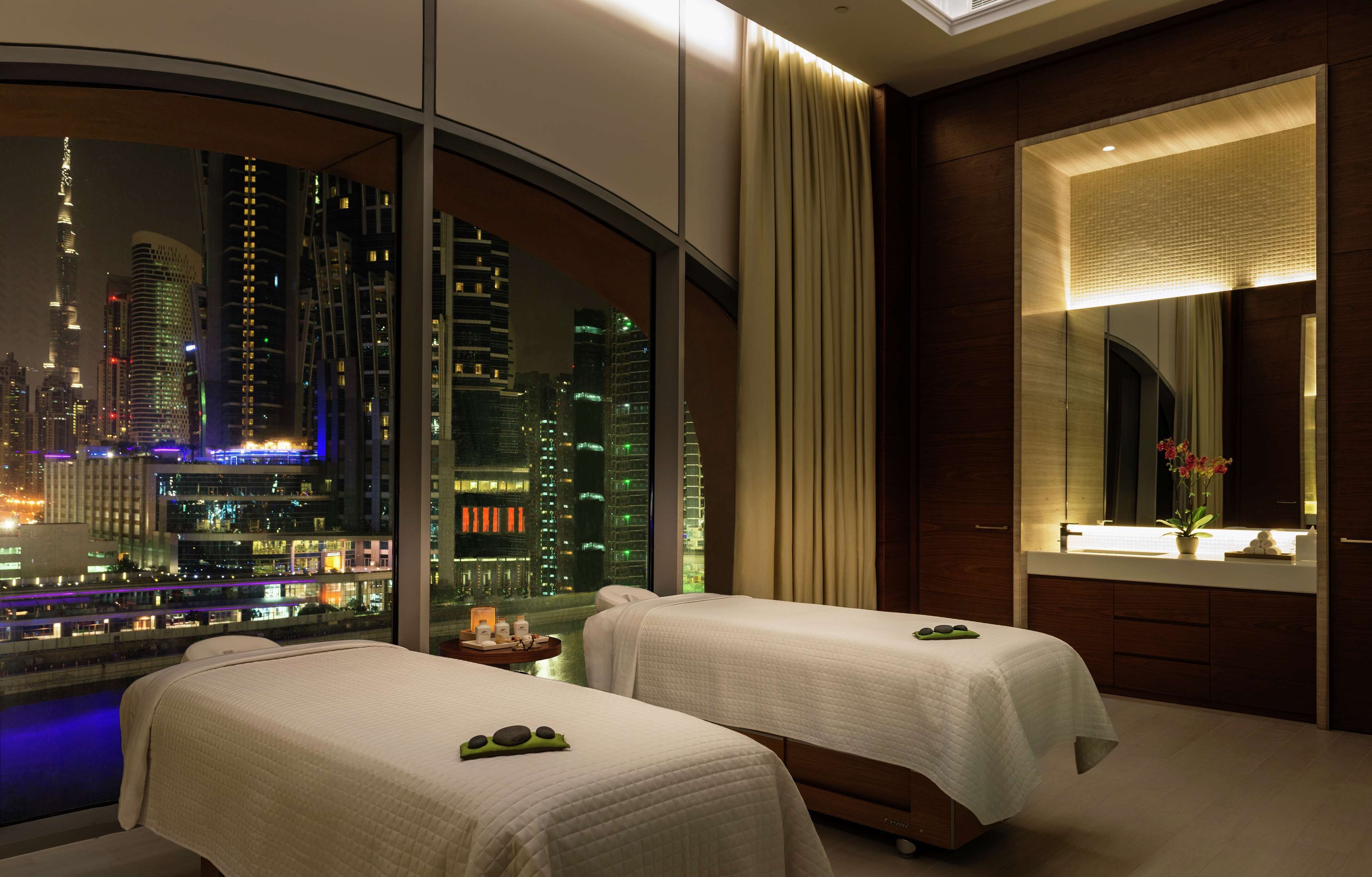 A hotel room with two beds, a large window with a city view, a bathroom with a mirror and sink, and a night sky visible through the window.