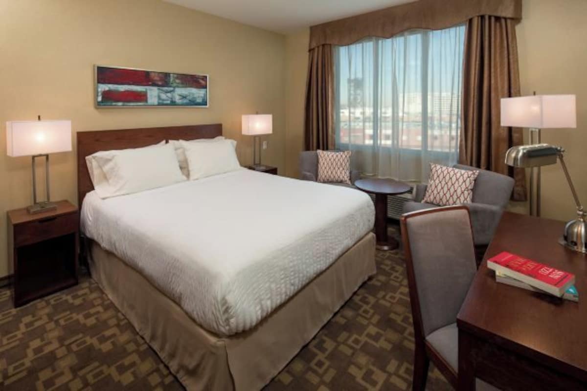 Habitación de hotel con una cama grande, dos mesitas de noche con lámparas, una mesa redonda pequeña con una silla y vistas a la ciudad a través de amplios ventanales.