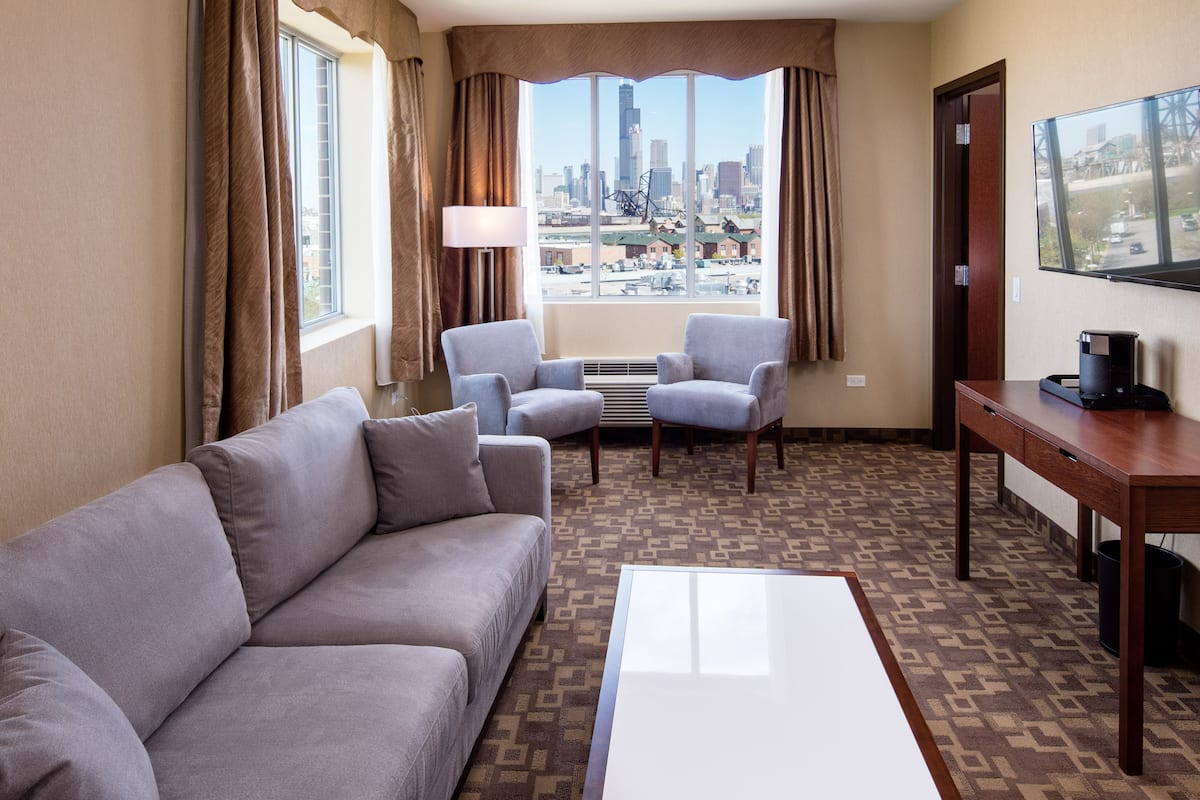 A hotel room with a sofa, two armchairs, a coffee table, a TV, and a city view.