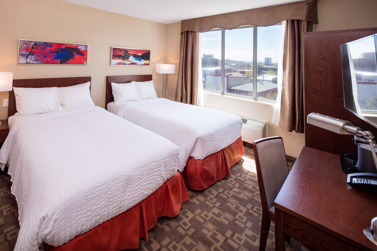 A hotel room with two beds, a desk, a chair, a TV, and large windows with a city view.