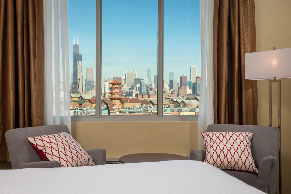 A hotel room with a large window offering a city view, two armchairs, a bed, and a lamp.