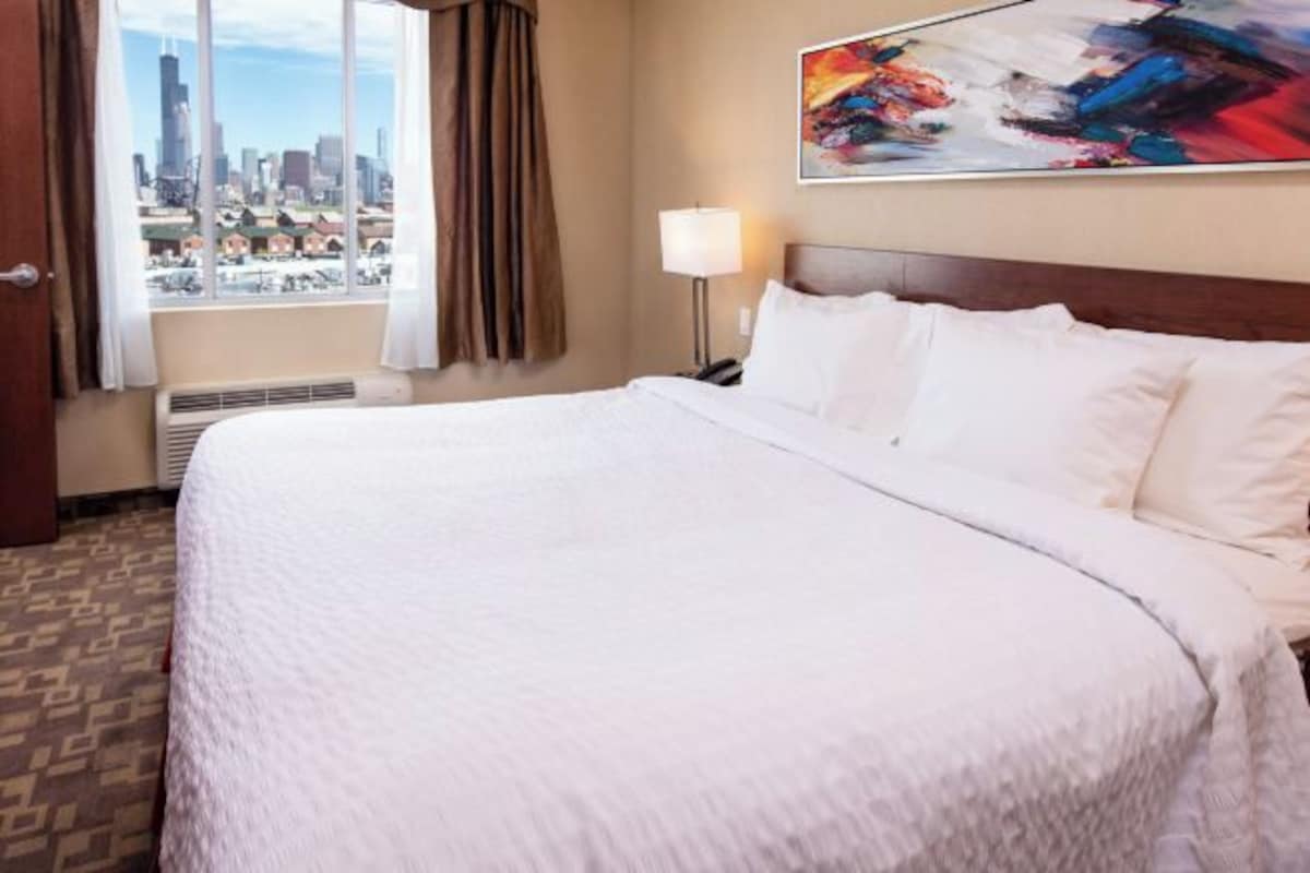 Habitación de hotel con una cama grande, vista a la ciudad a través de la ventana, un cuadro en la pared y una mesita de noche con una lámpara.
