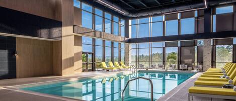An indoor swimming pool with multiple sun loungers, a tiled floor, and large windows.
