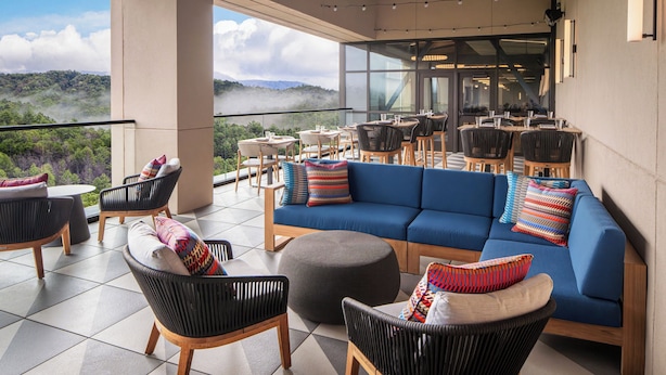 A modern outdoor seating area with a blue sofa, black and wooden chairs, and a view of a mountainous landscape. - Harrah's Cherokee Valley River Hotel & Casino – A Caesars Rewards Destination (Murphy)