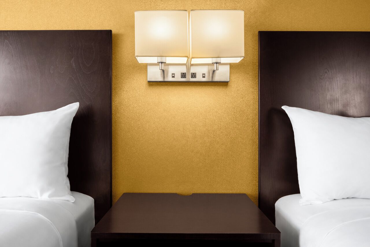 Habitación de hotel con dos camas, una mesita de noche y una lámpara de pared entre ellas.