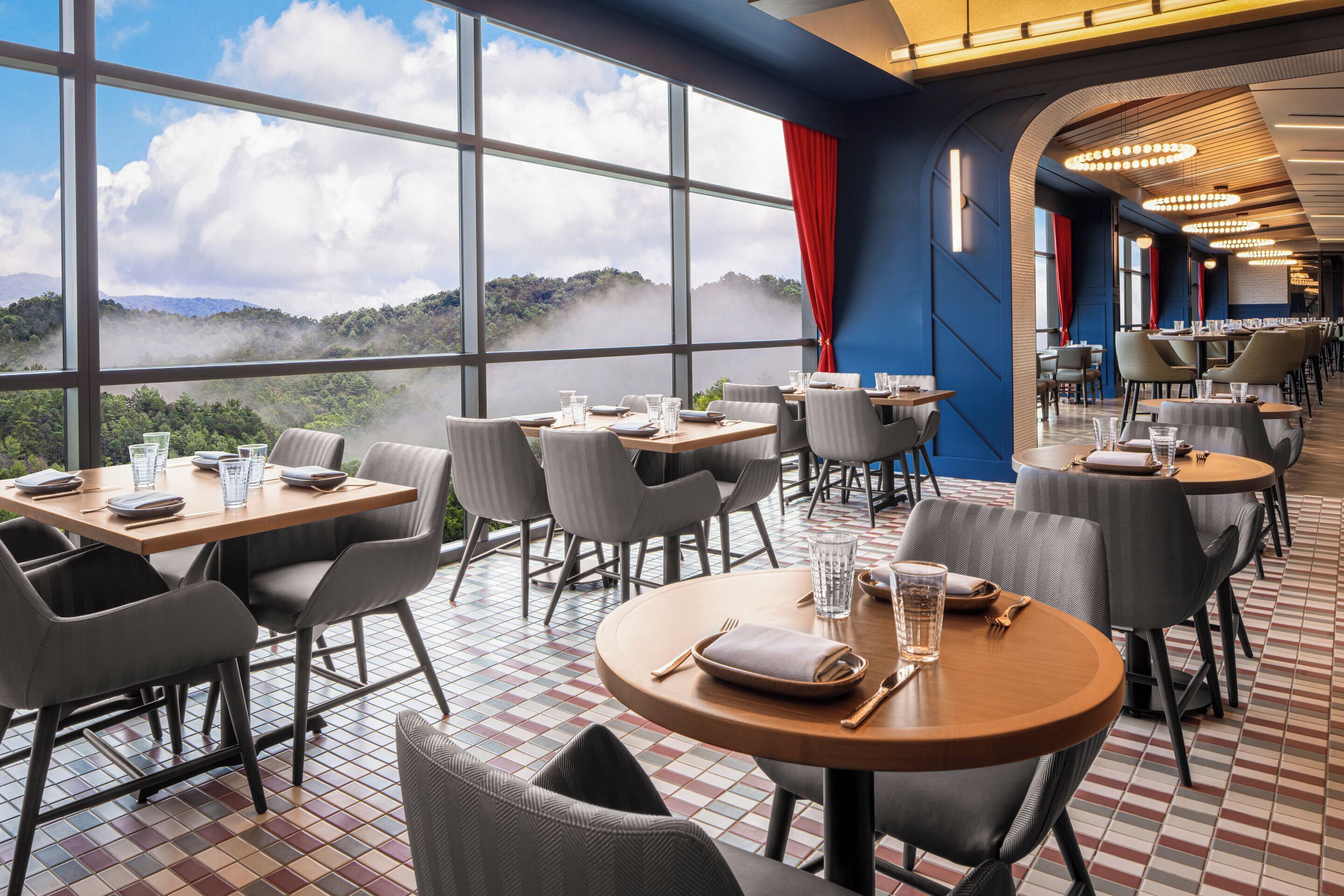 A restaurant with tables and chairs arranged for dining, large windows offering a view of a mountainous landscape, and a patterned floor.