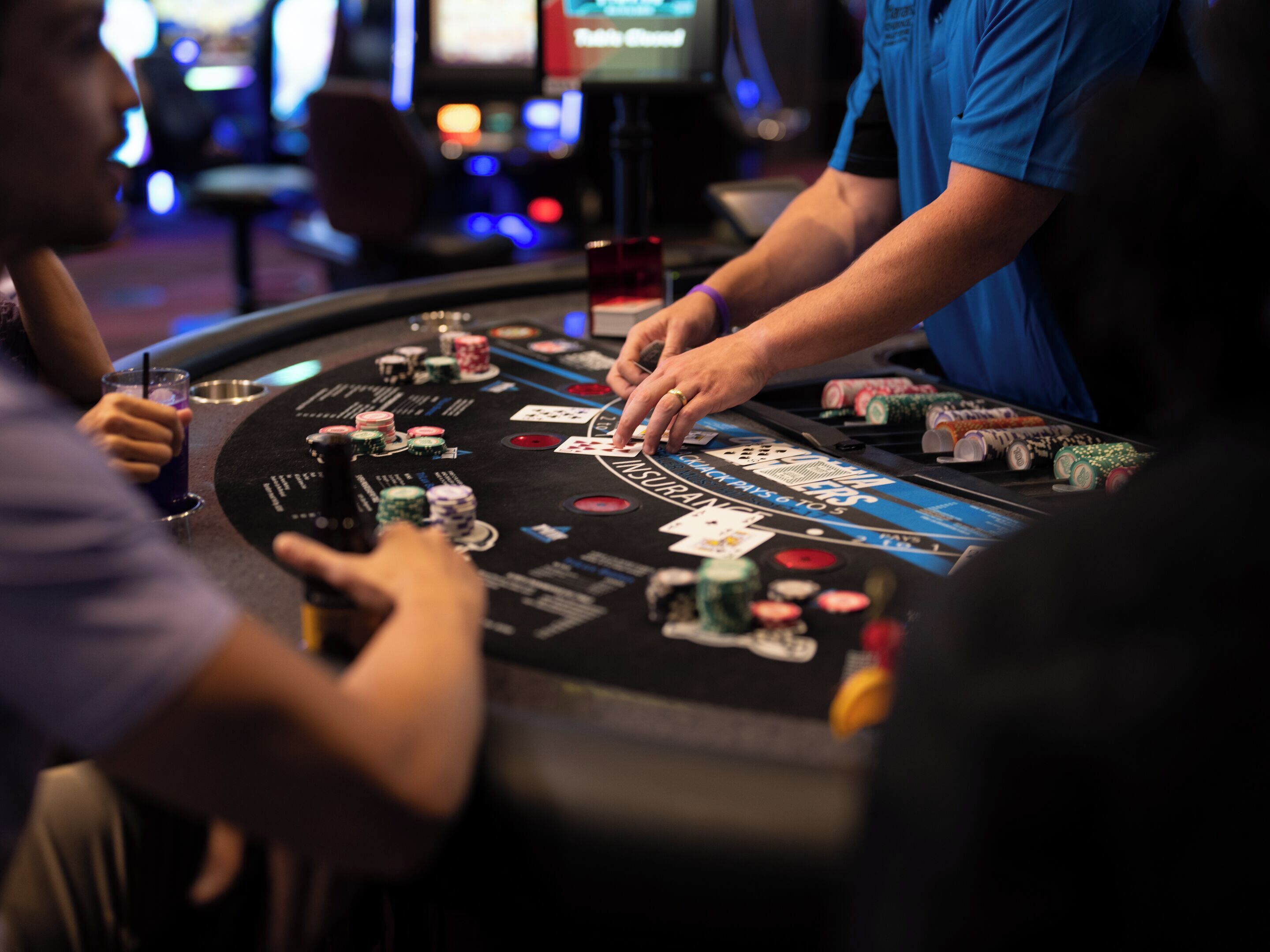 A casino table with chips and cards, a person dealing cards, and a blurred background with a screen.
