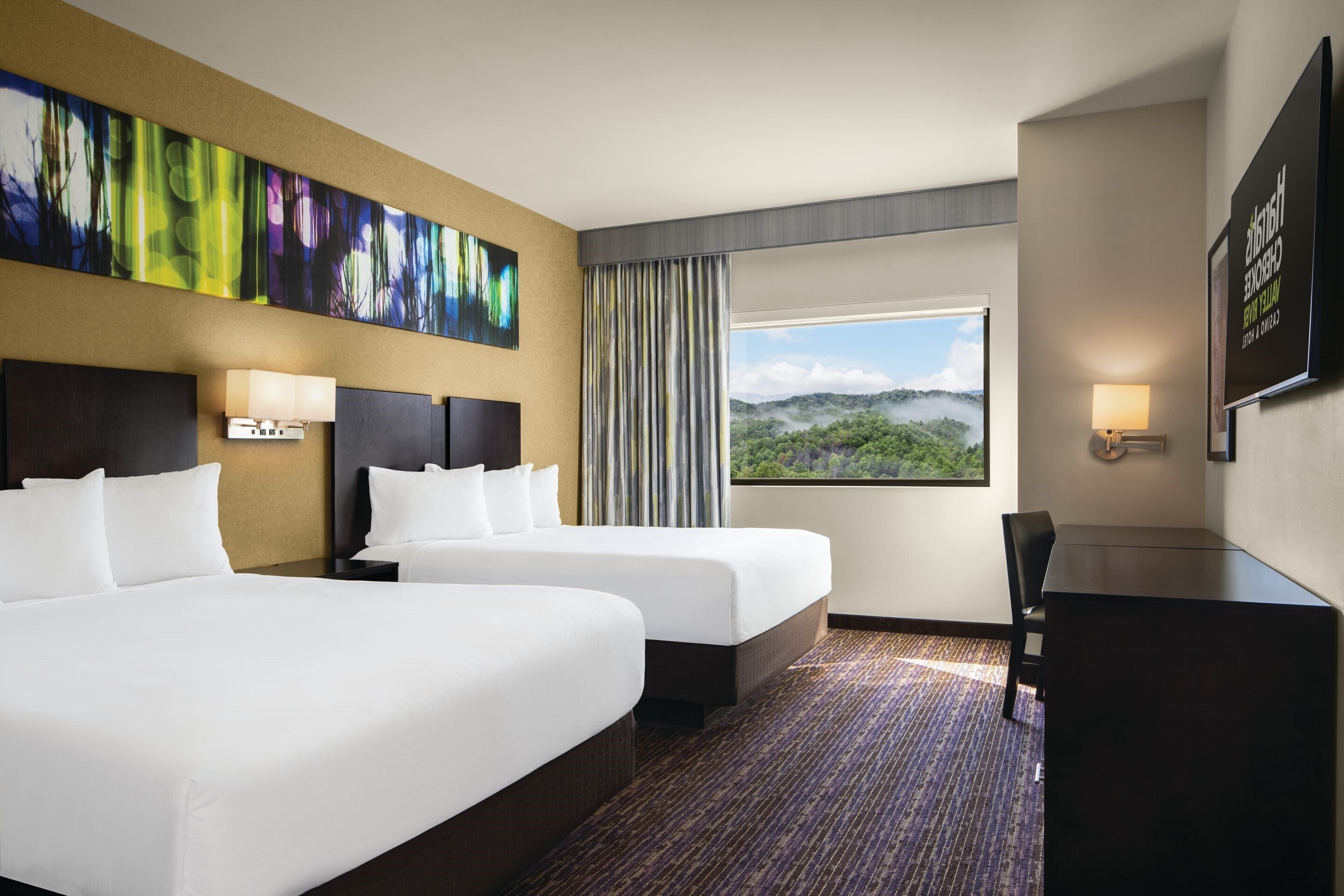 Quarto de hotel com duas camas, uma escrivaninha, uma janela grande com vista para o verde e um quadro na parede.
