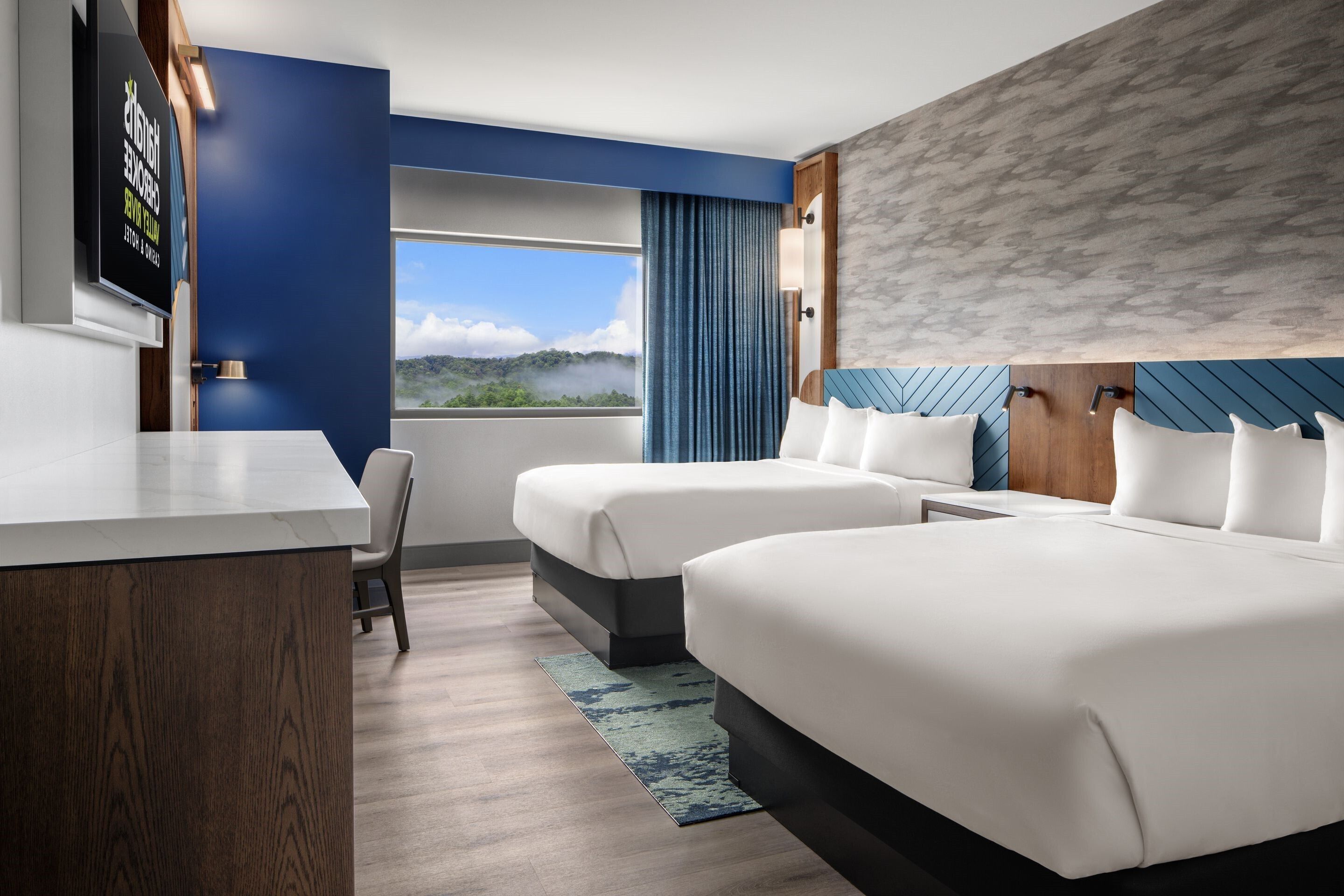A hotel room with two beds, a desk, a TV, and a view of a mountainous landscape.