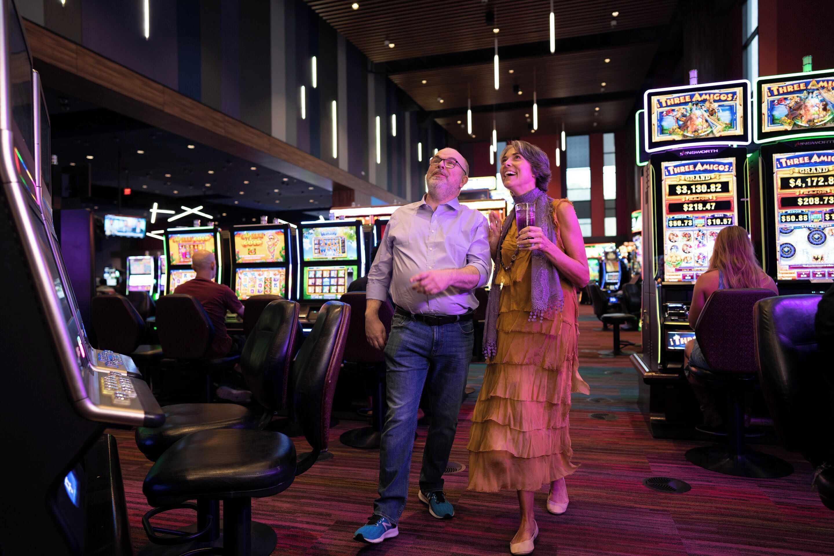 Een casino interieur met gokkasten, een man en een vrouw staan er, en een bar.