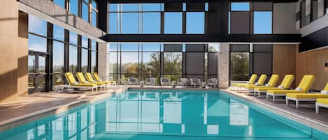 Una piscina interior moderna con paredes de vidrio transparente, sillas de descanso amarillas y vistas a árboles.