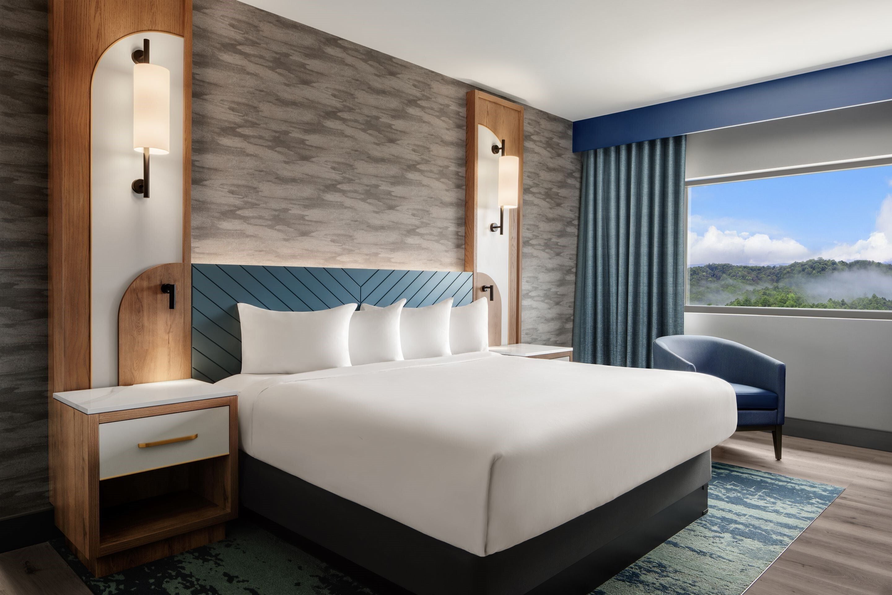 A modern hotel room with a large bed, a bedside table, a chair, and a view of the outdoors.