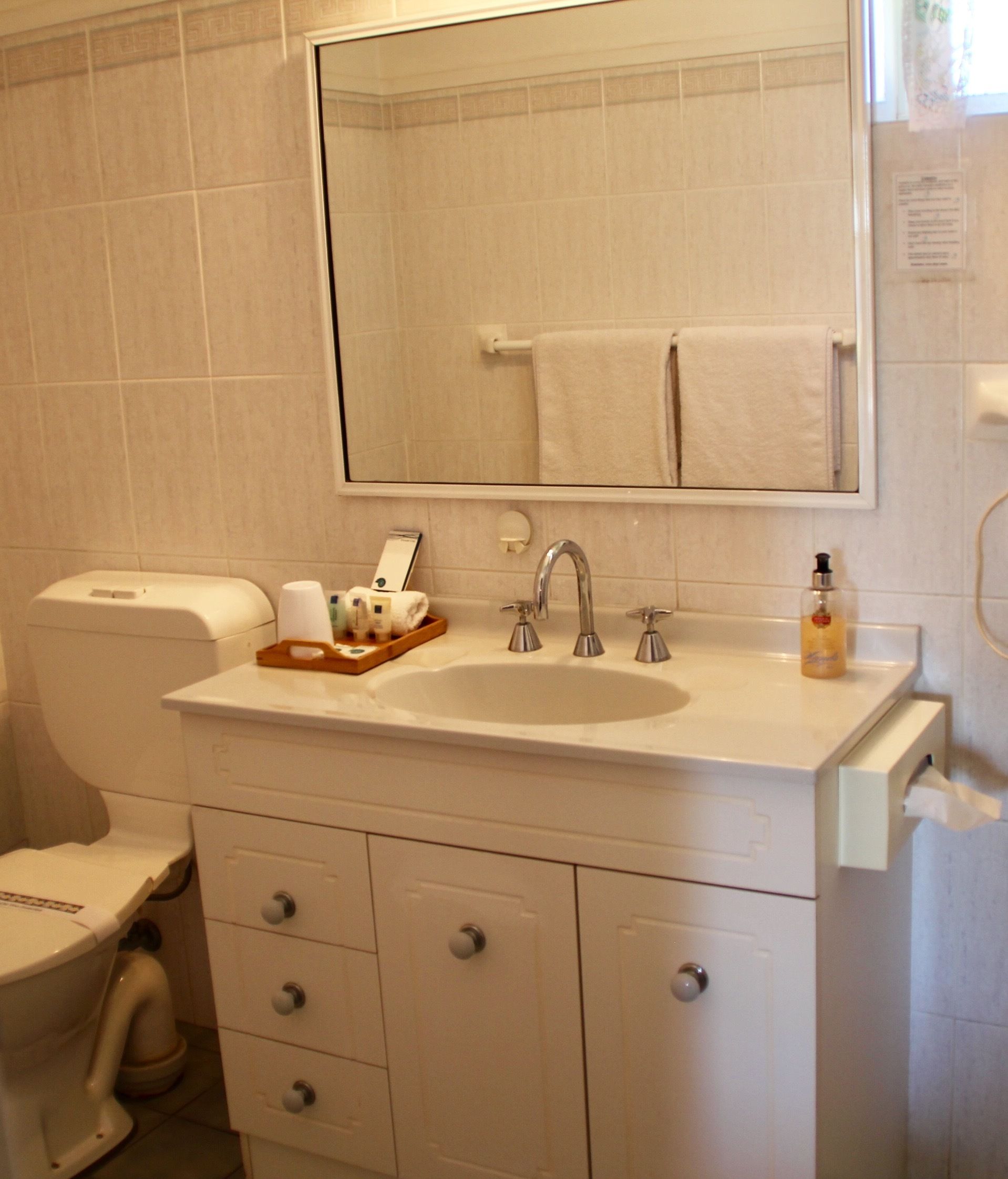 A bathroom with a white sink, two faucets, a mirror, a toilet, and a trash bin.