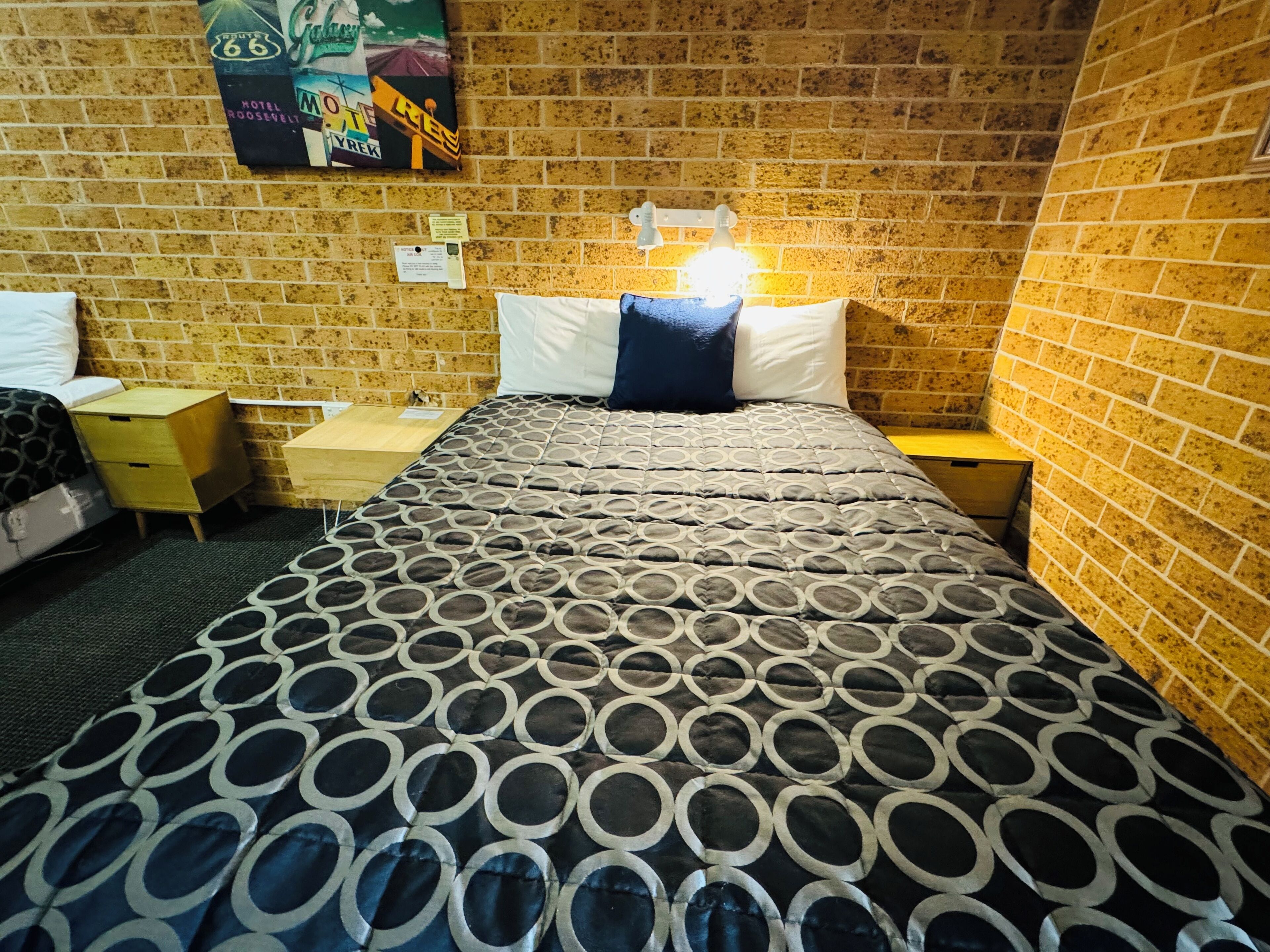 A hotel room with a brick wall, a bed with a patterned bedspread, a nightstand, a bedside table with a lamp, and a poster on the wall.