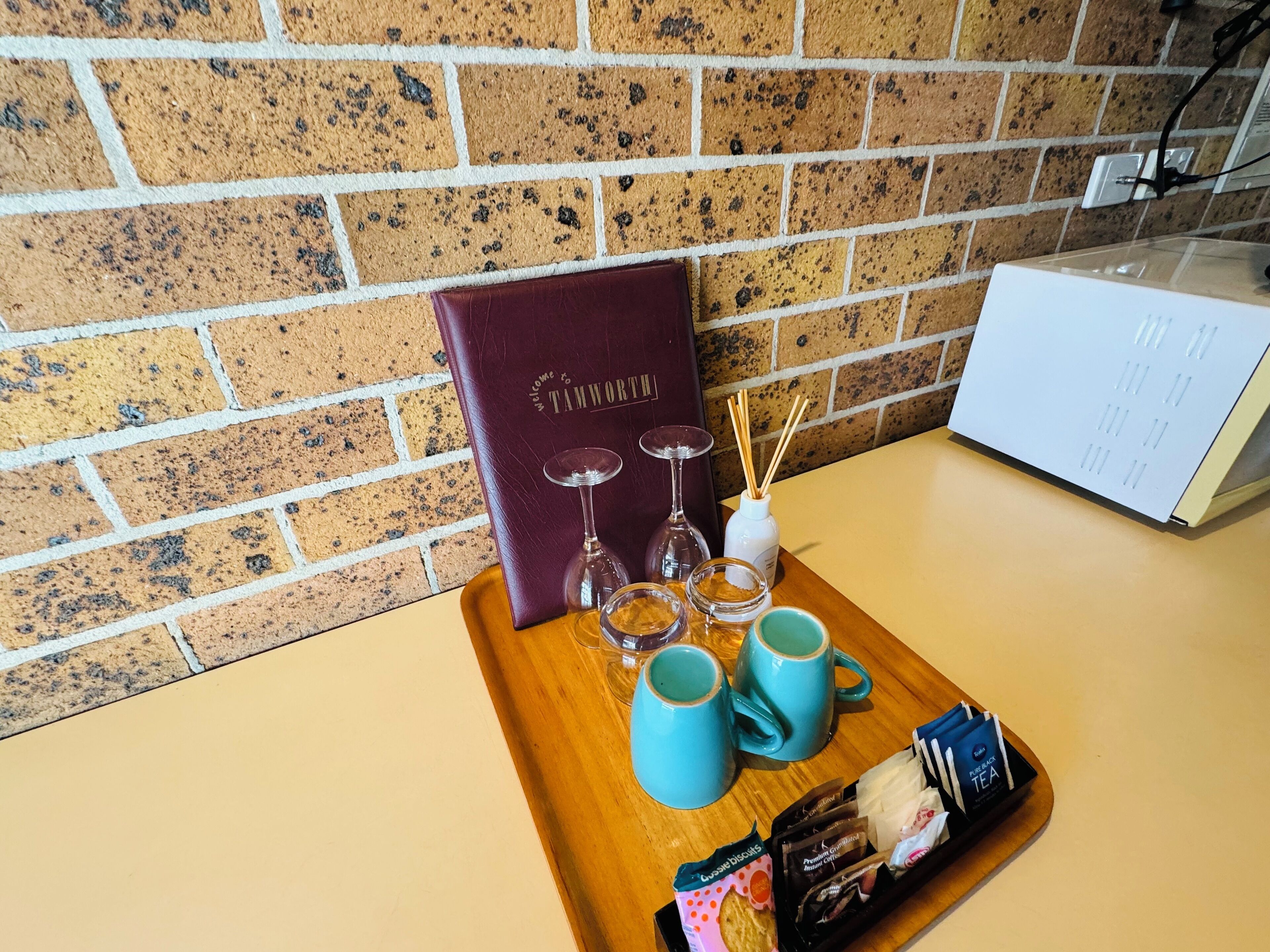 Una bandeja con un libro de bienvenida, copas de vino, una botella de cañas de jarrón, dos tazas azul verdoso y paquetes de té y galletas.