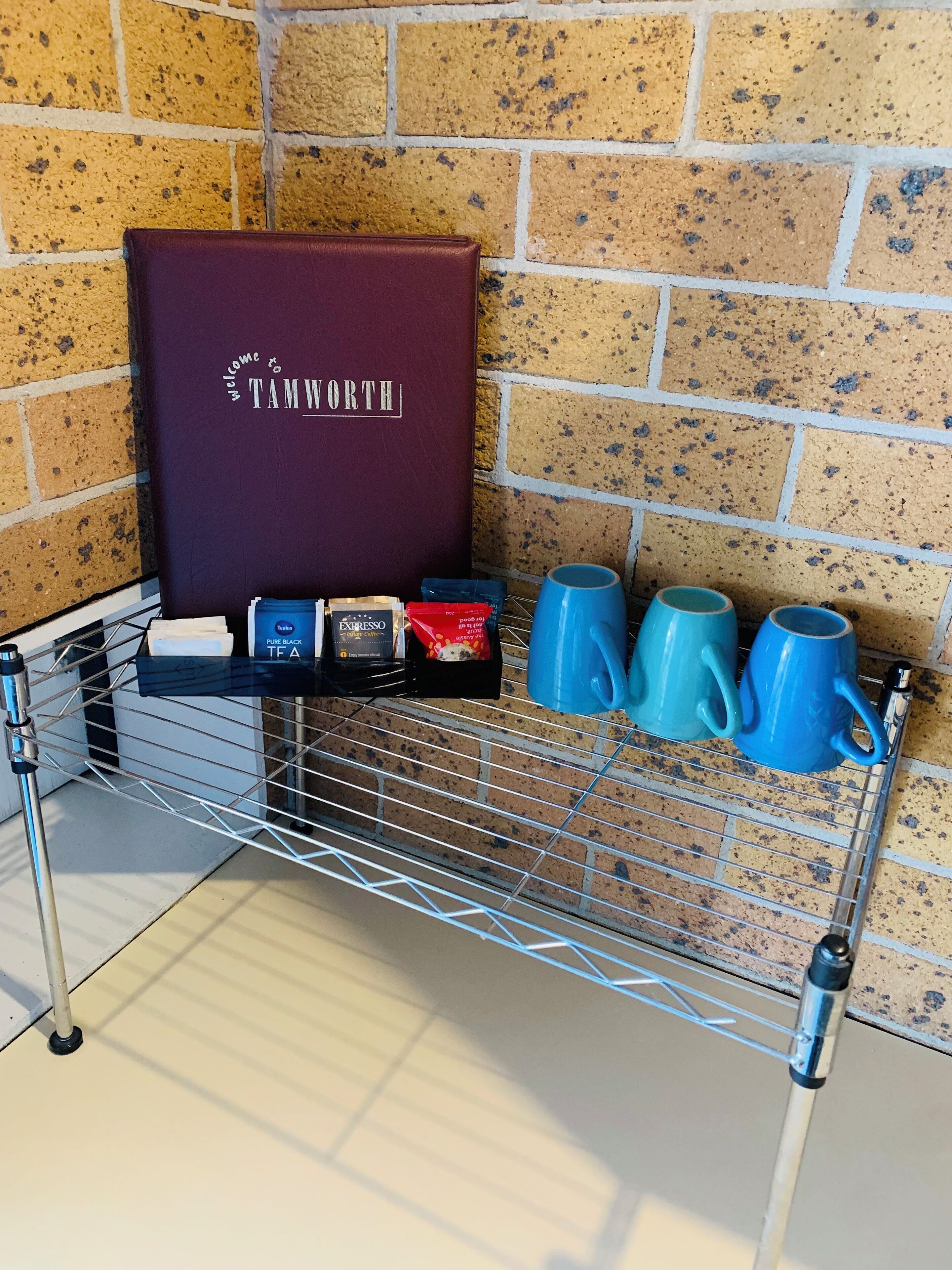 A metal rack with a welcome sign, coffee bags, and blue mugs against a brick wall.