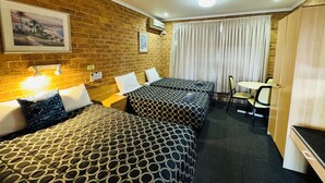 Ein Hotelzimmer mit zwei Betten, einem Schreibtisch und einem Stuhl. Es gibt zwei Wandlampen, ein Fenster mit Vorhängen und eine Ziegelwand.